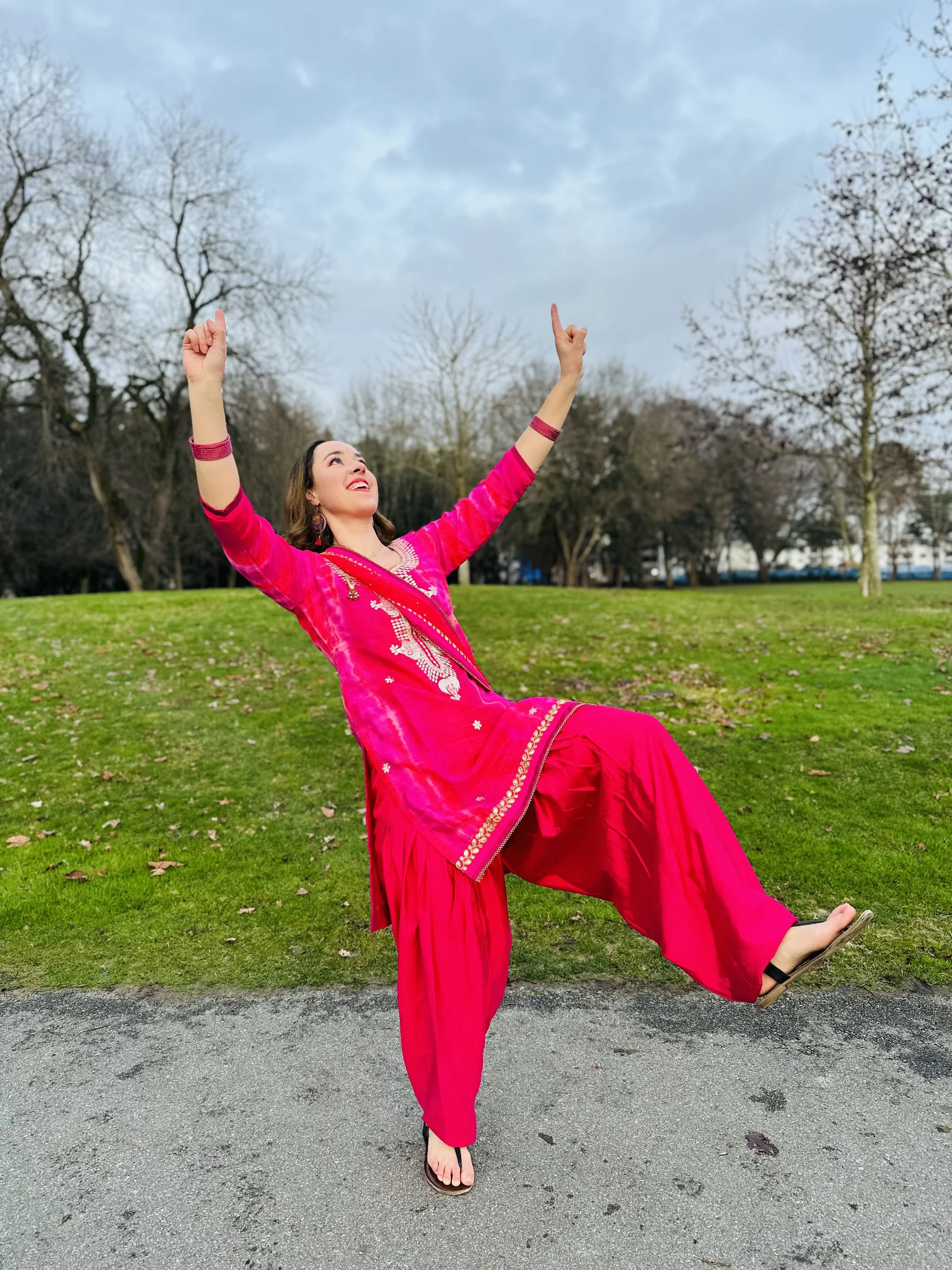 A woman in a bright pink traditional Punjabi outfit joyfully doing Bhangra outdoors on a cloudy day.