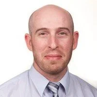 Headshot of a bald man with a light complexion, wearing a white dress shirt and a striped tie, smiling against a white background.
