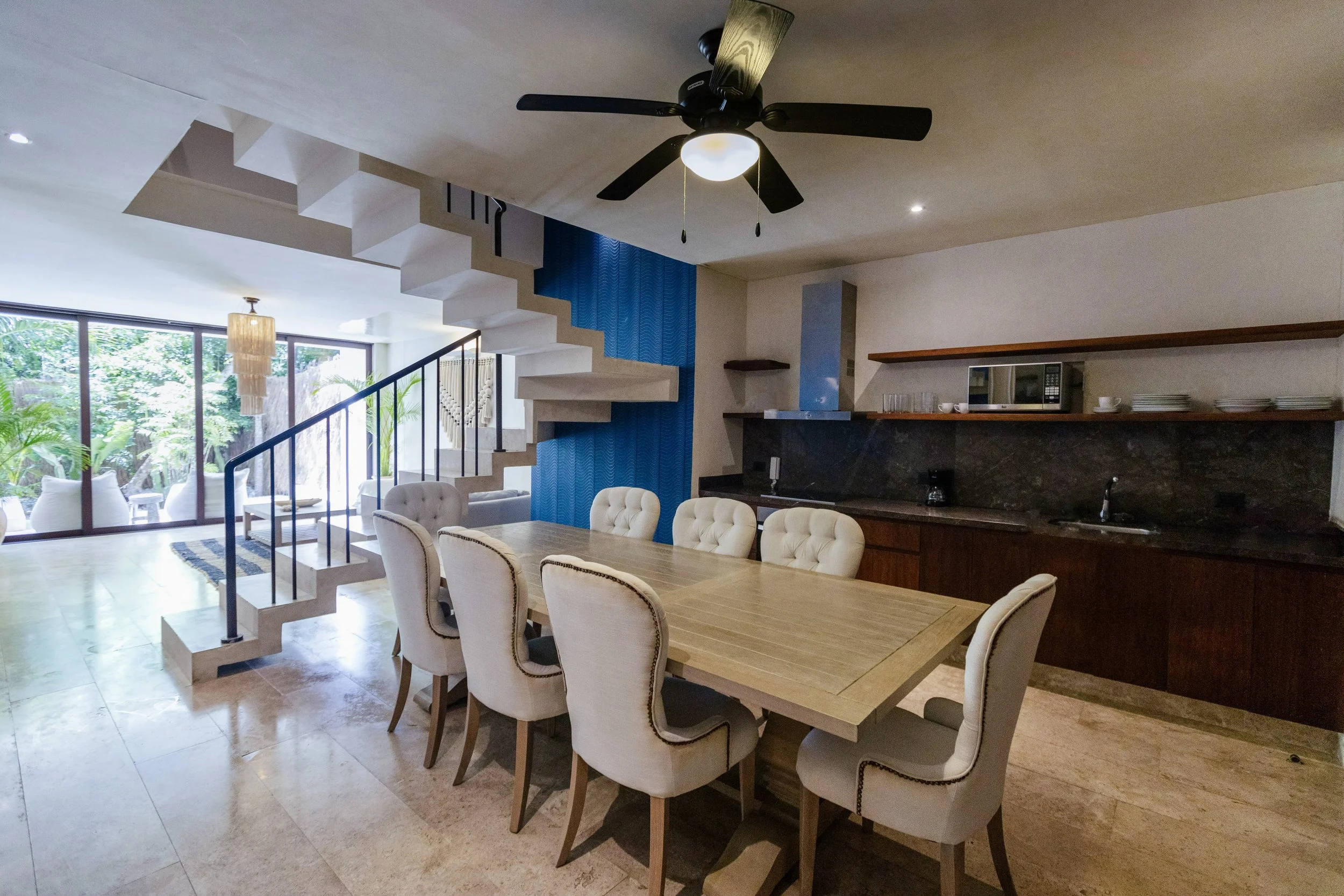 Interior of a modern dining room with a large wooden table, upholstered beige chairs, a black ceiling fan, a kitchen area with black countertops, a microwave, and open shelves, and a staircase leading to an upper level, with large windows showing a patio and greenery outside.