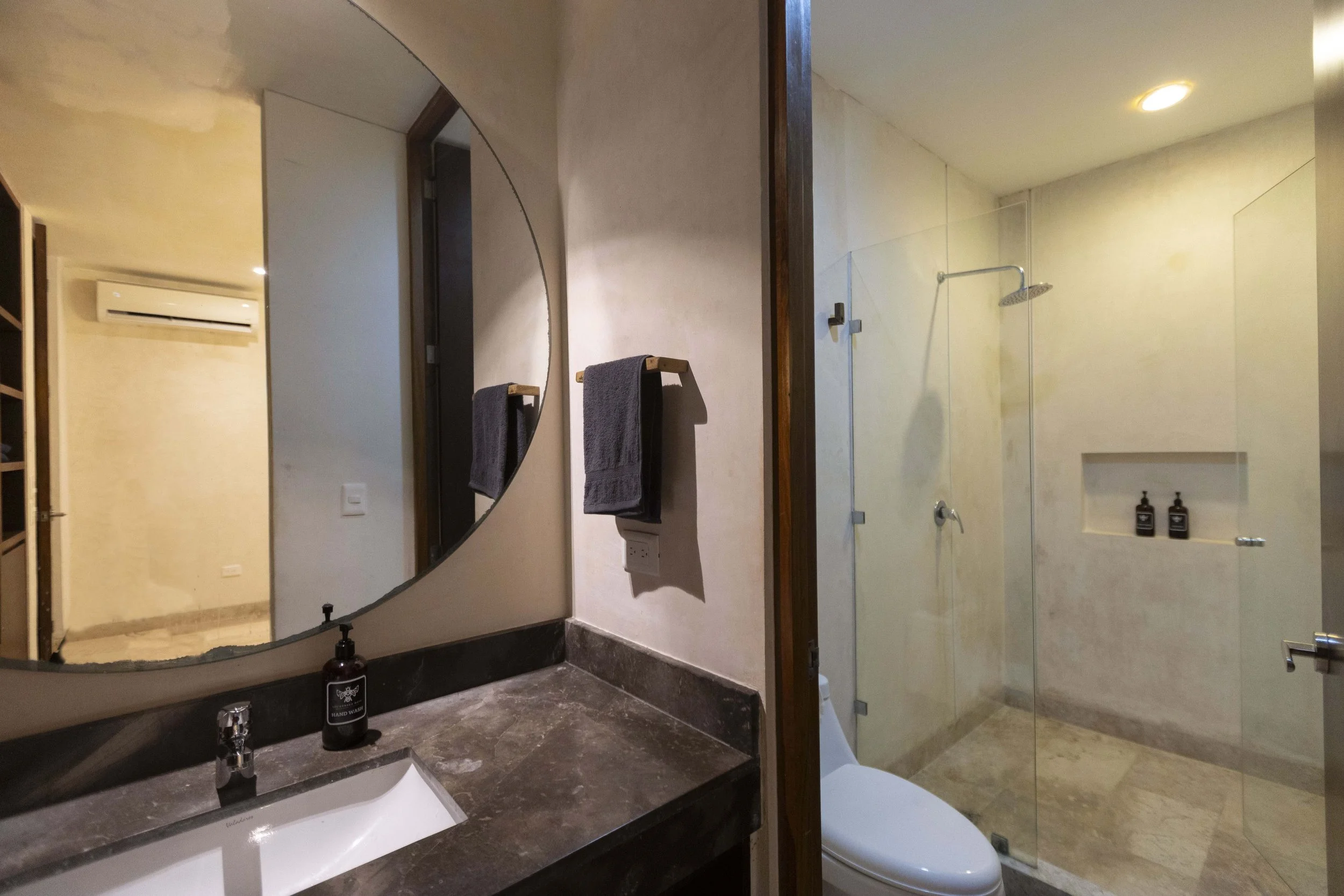 Bathroom with a large round mirror, a black countertop with a sink and soap dispenser, and a separate shower area enclosed with glass. Towel hanging on a wall hook.
