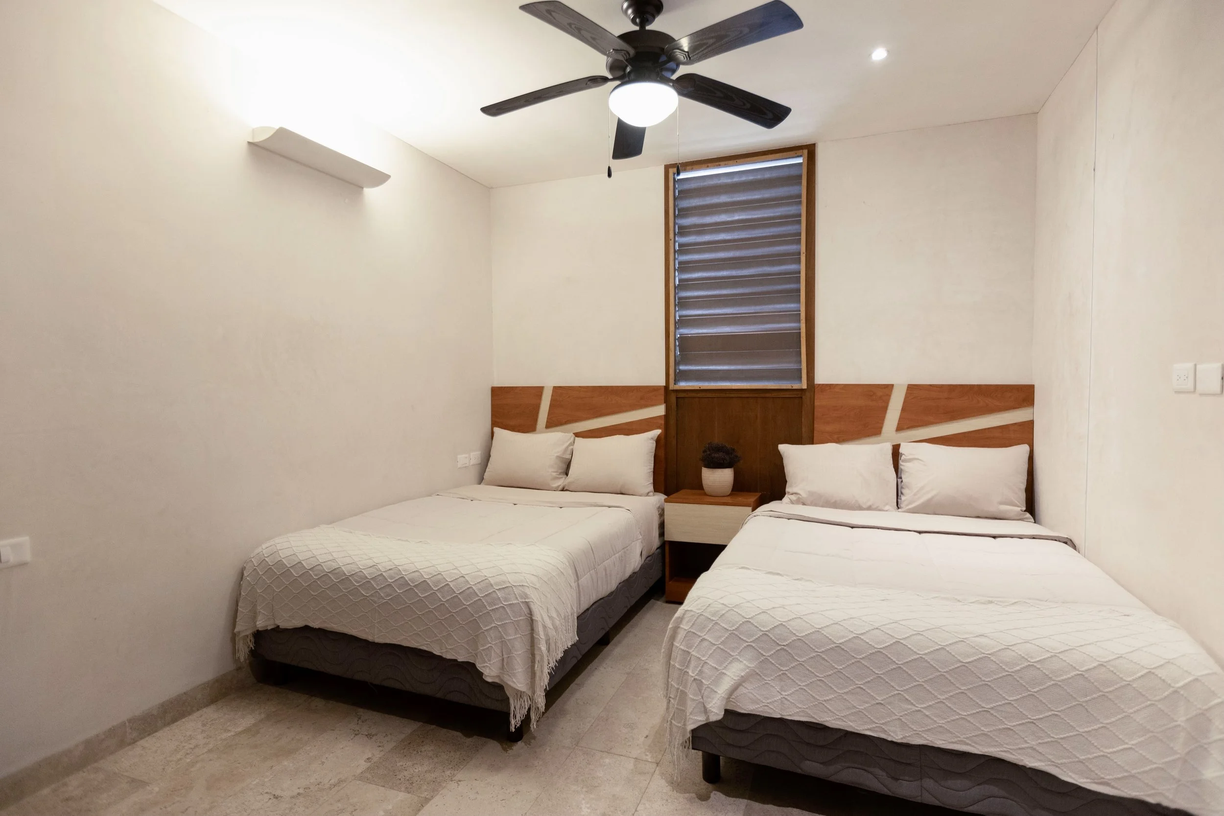 Two beds with white bedding and pillows in a minimalist bedroom, wooden headboards, a nightstand with a plant, a window with blinds, a ceiling fan, white walls, and marble floors.