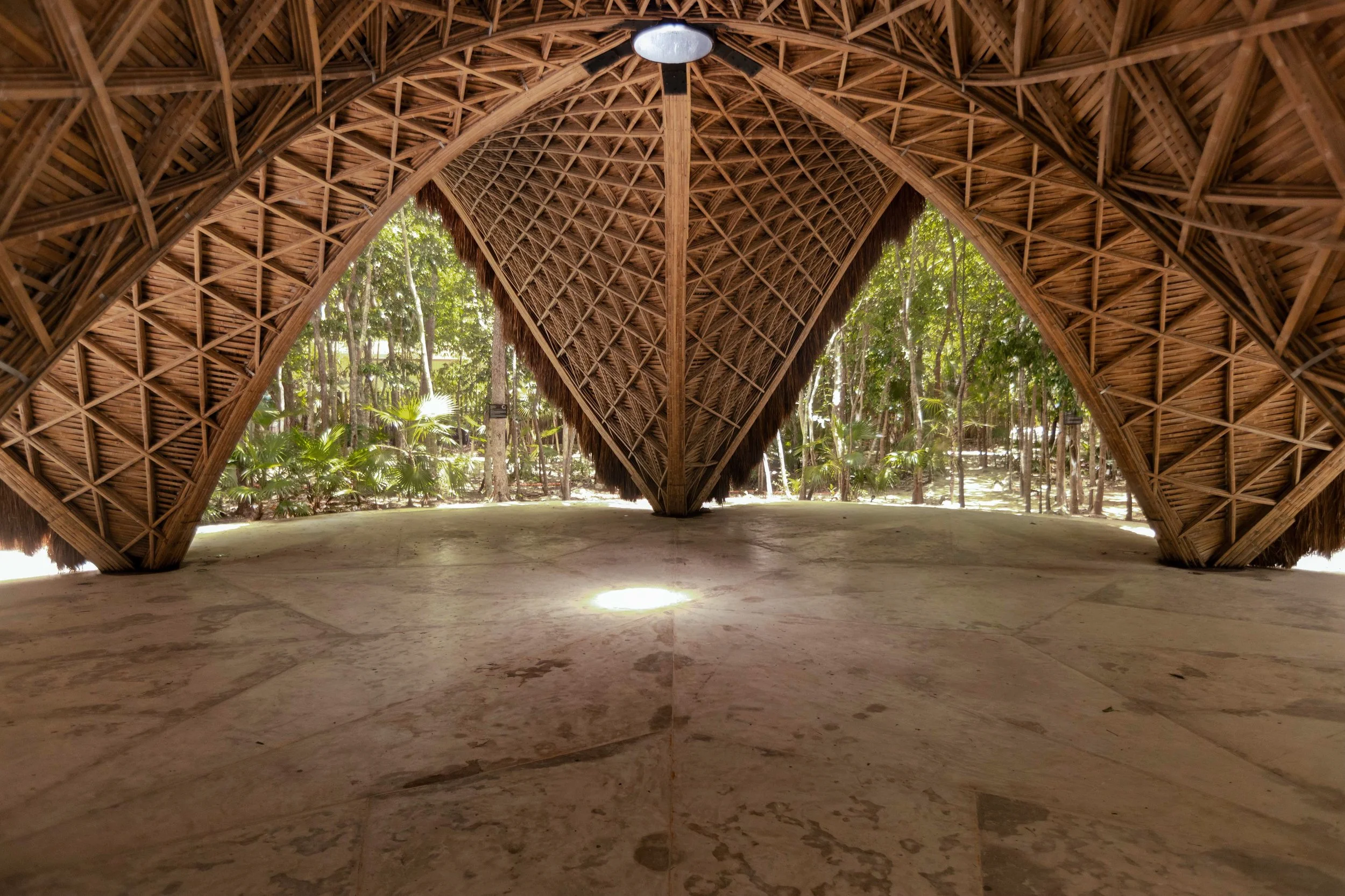 Interior of a thatched wooden structure with open sides, surrounded by lush green trees and plants, with a circular stone floor.