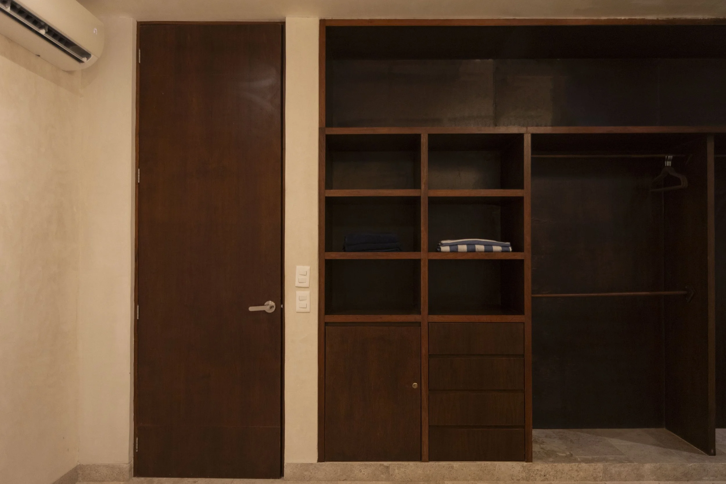 Empty wooden closet with shelves and hanging space, two folded towels or cloths on one shelf, and a dark door next to it in a room with beige walls.