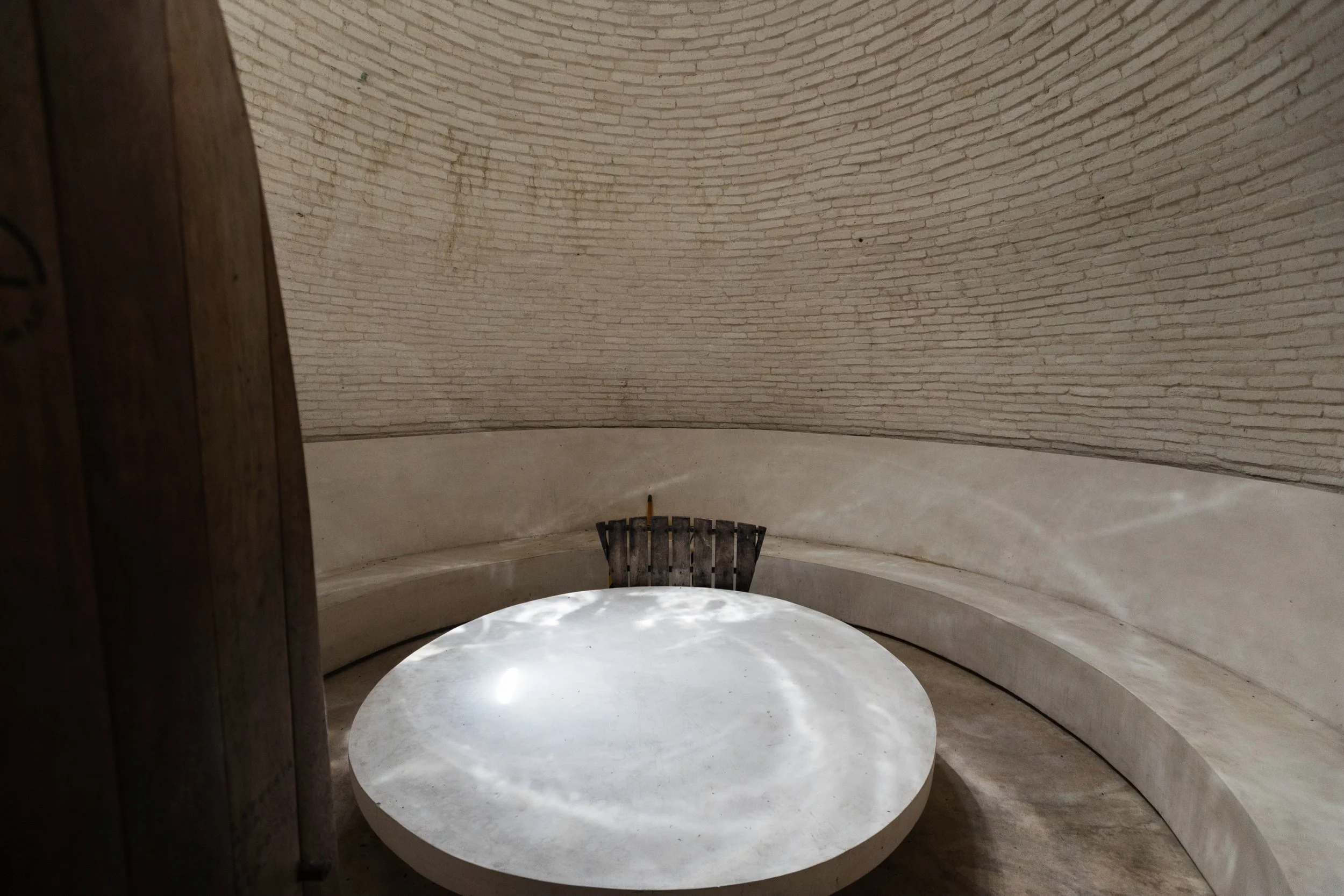 A round white marble table in a semi-circular corner with a curved brick wall and a bench along the back wall. A small black wooden bench is at the end of the curved bench. The space appears to be indoors with natural lighting.