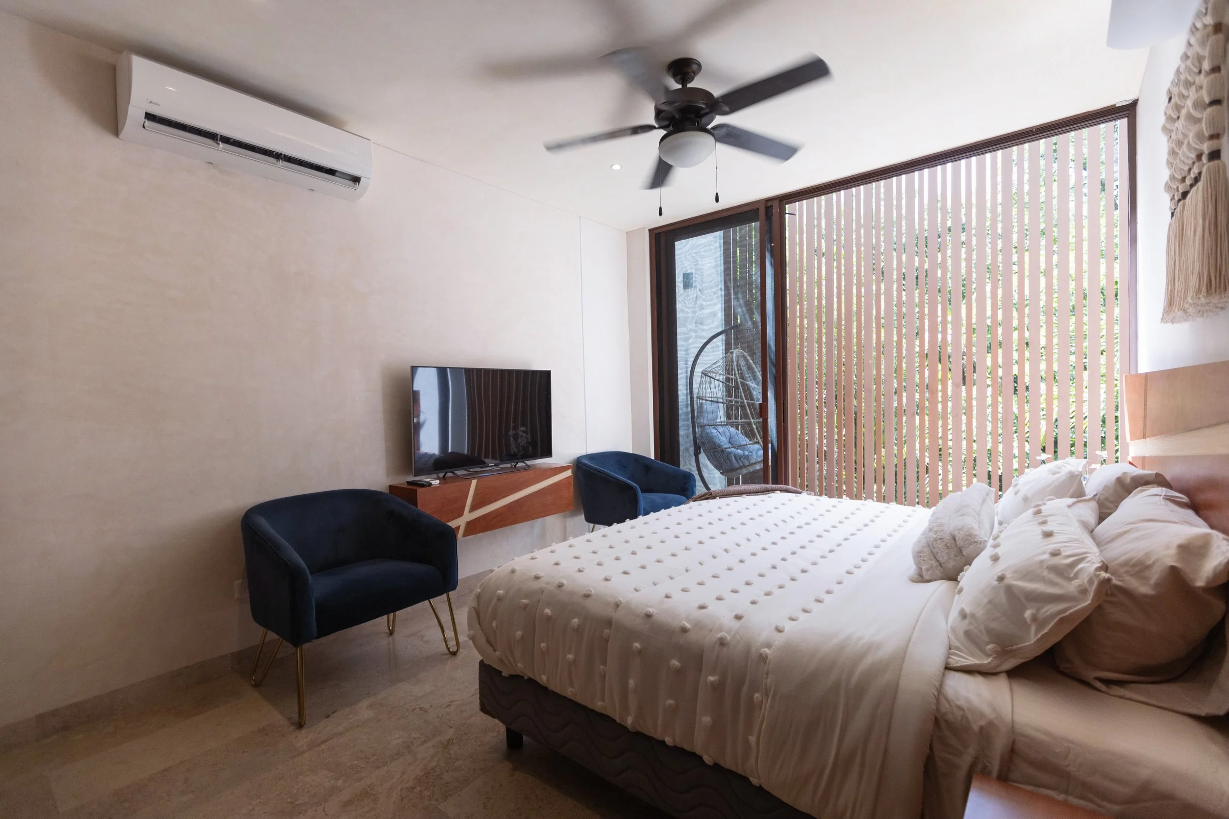 A bedroom featuring a white bed with multiple pillows, a black ceiling fan, a wall-mounted air conditioner, a flat-screen TV on the wall with a wooden shelf, two navy blue chairs, and a hanging chair outside through large glass sliding doors with wooden slats.