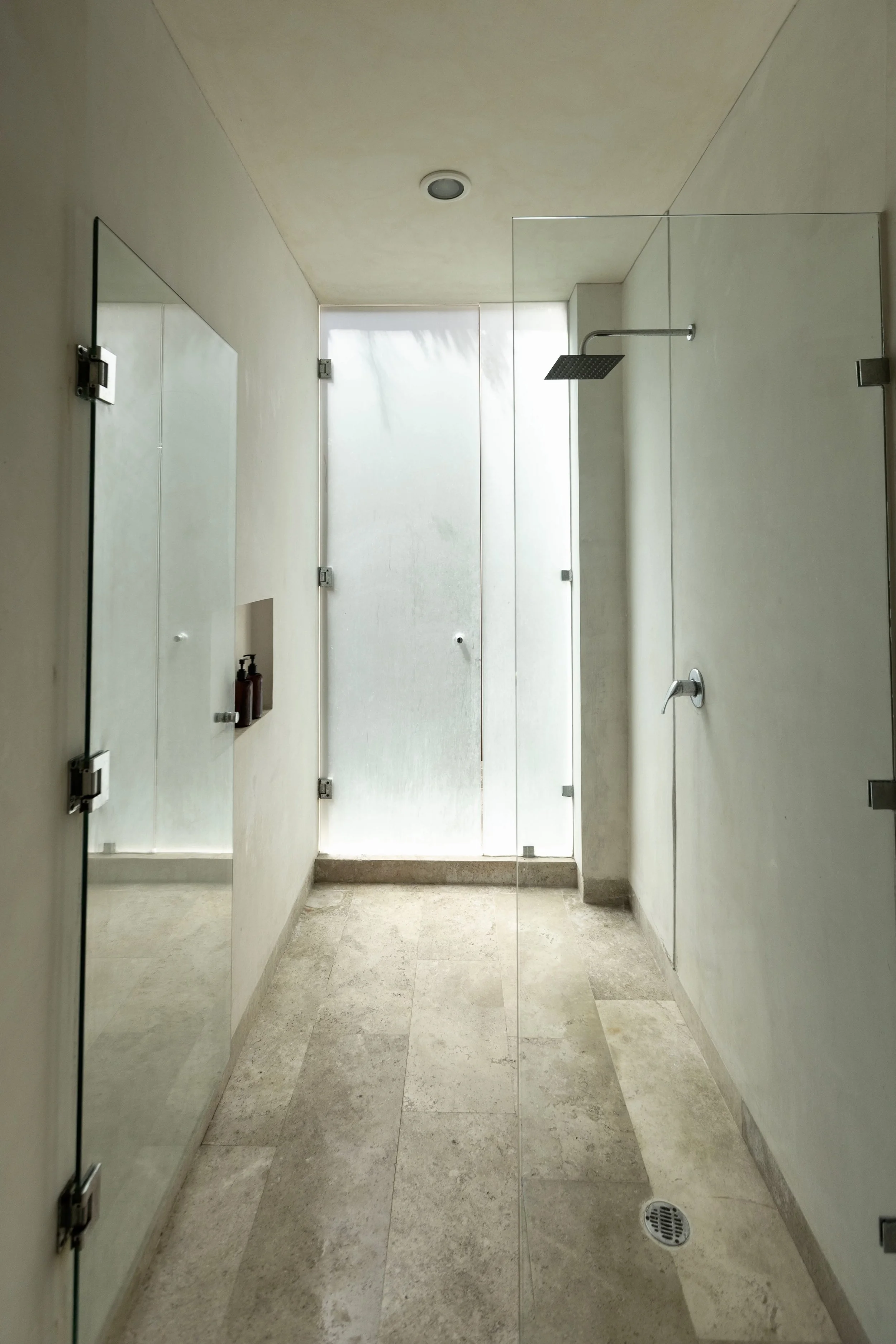 Minimalist walk-in shower with frosted glass door, rain showerhead, and built-in niche with soap and shampoo bottles.