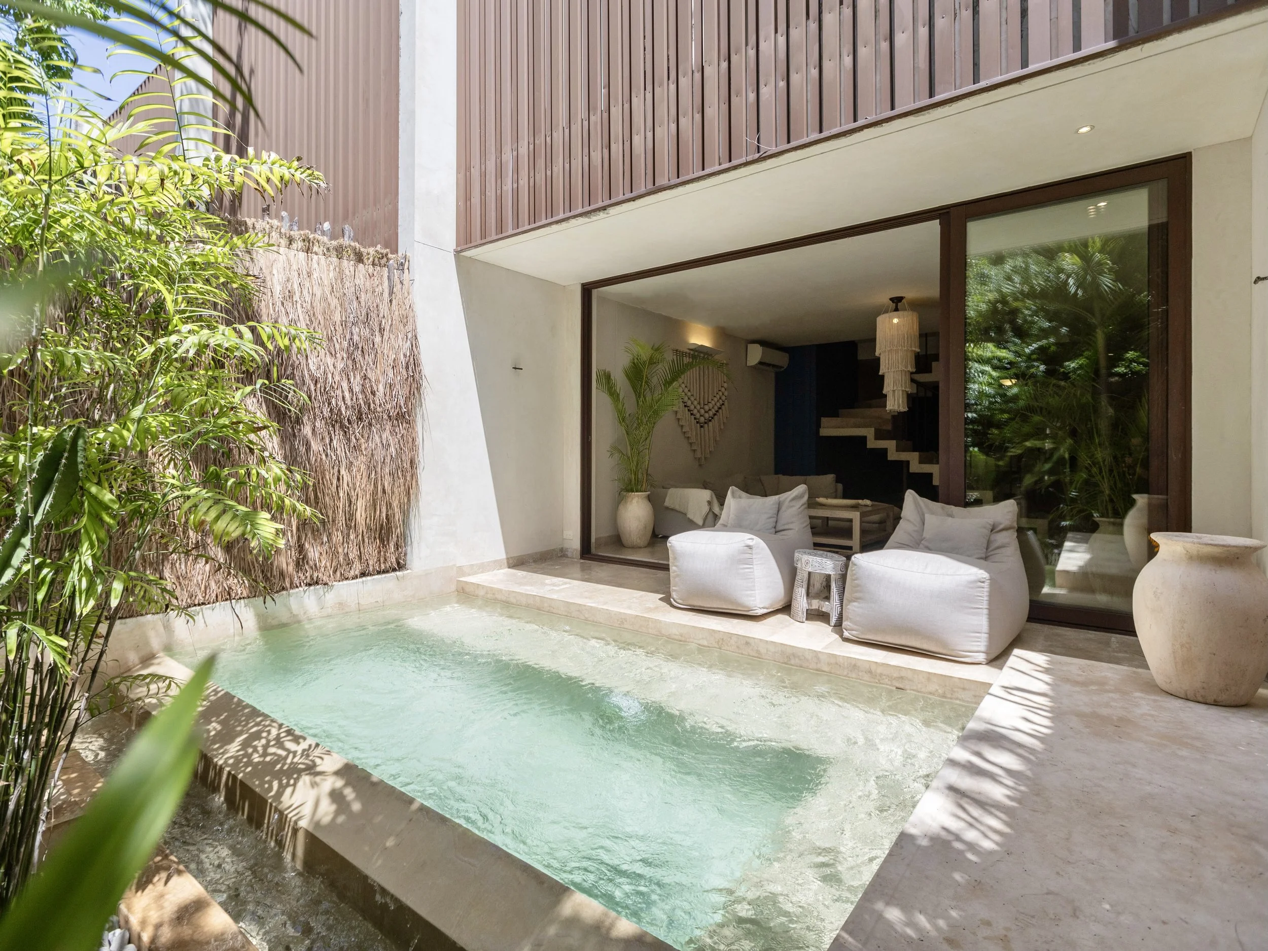 Modern indoor living area with large glass sliding door, white bean bag chairs, potted plants, and a small pool outside surrounded by greenery.