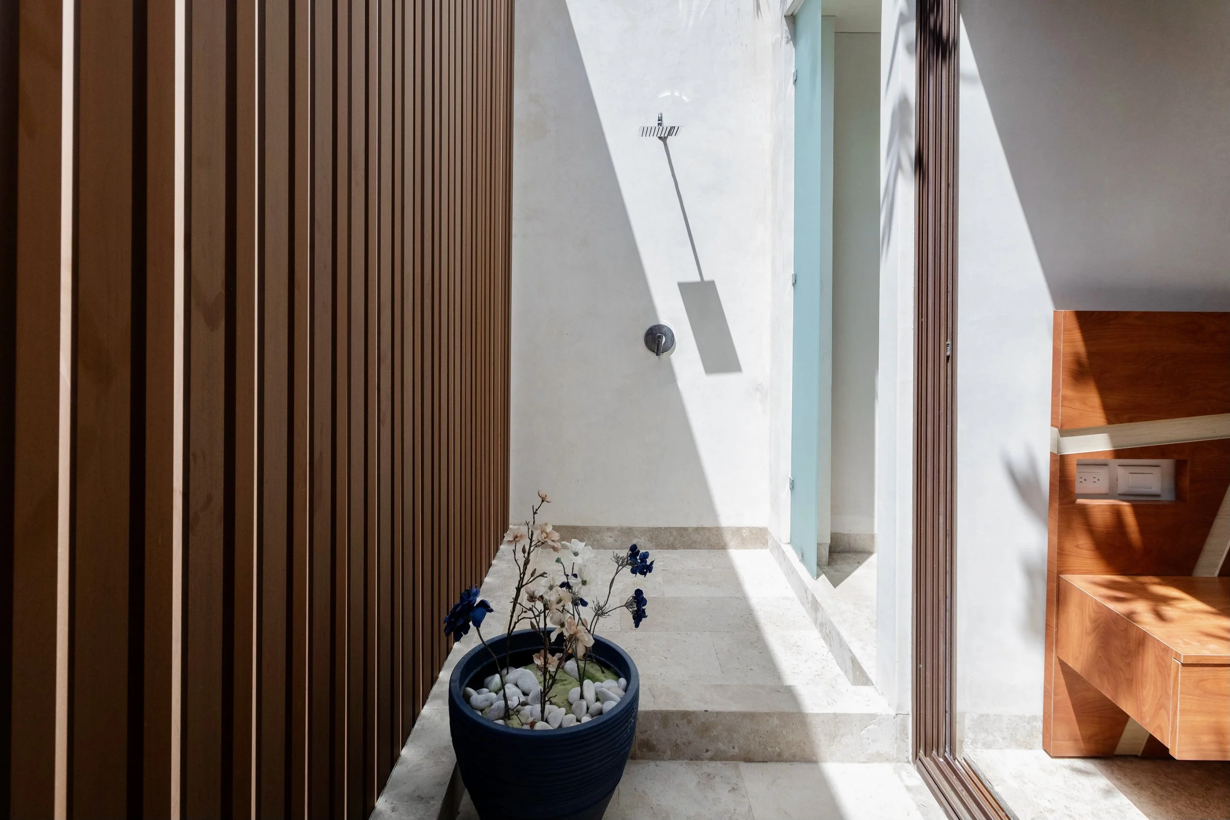 outdoor space with wooden slat wall on the left, a white wall with outdoor shower and shower head, a potted plant with white and blue flowers and white stones, and a wooden piece of furniture on the right side. Sunlight casts shadows in the space.