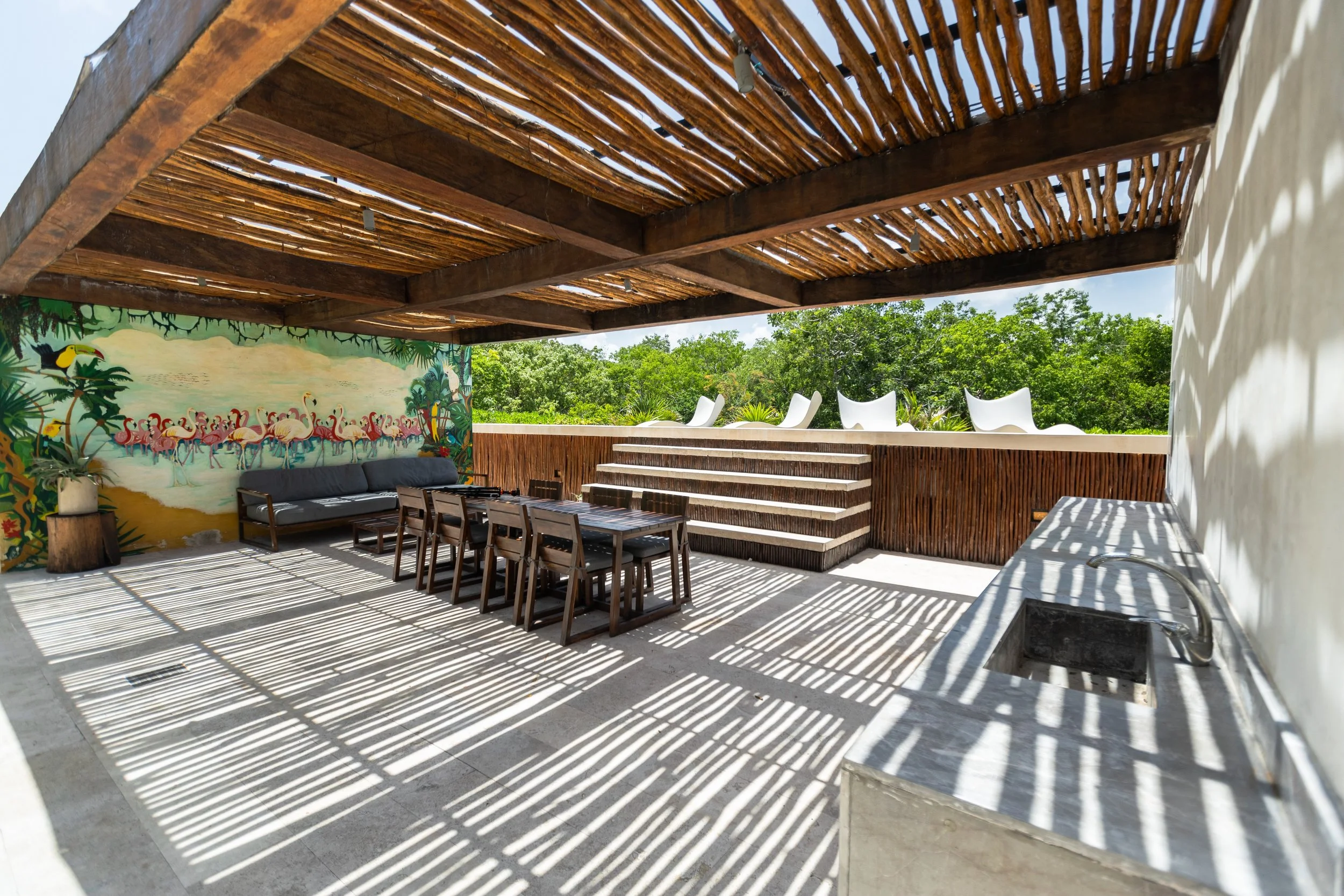 Outdoor patio with a wooden pergola casting striped shadows, a mural of flamingos and jungle scene on the wall, a long table with chairs, a black sofa, white lounge chairs on a raised platform, greenery in the background, and a sink with a faucet.