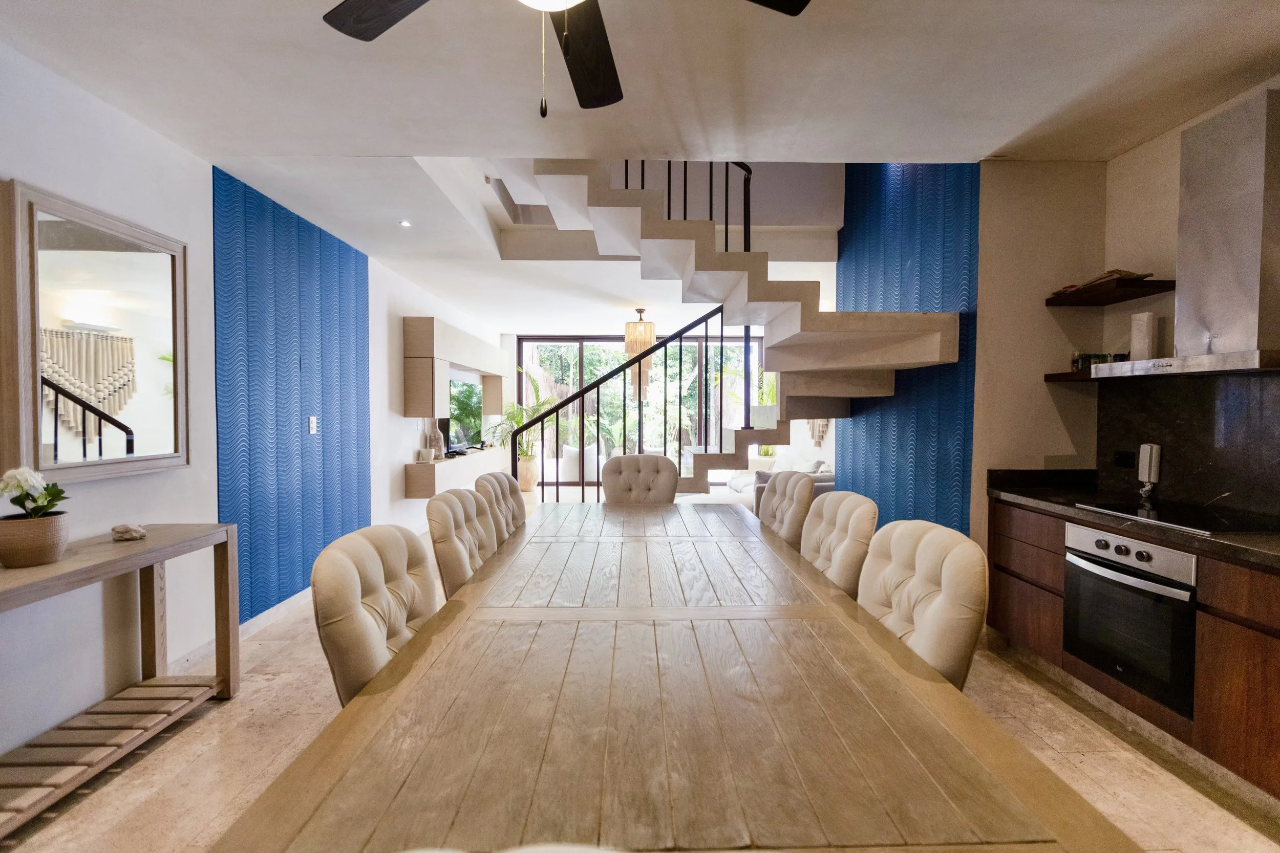 Dining area with a long wooden table, tufted beige chairs, blue textured walls, staircase, and large window with outside greenery.