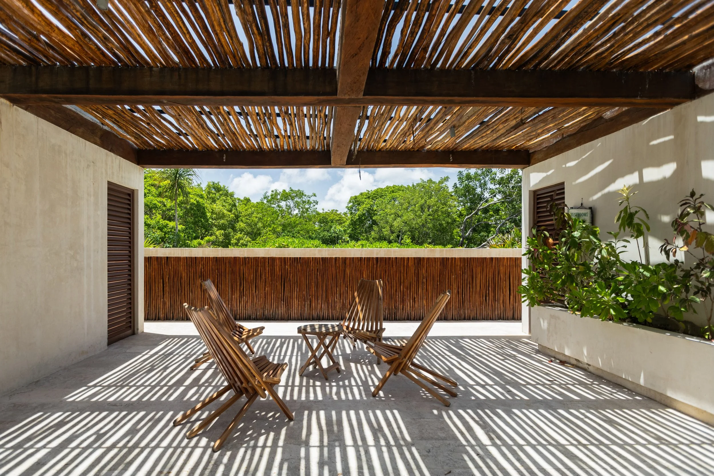 Sunlit balcony with wooden furniture and bamboo roof, overlooking lush green trees with partly cloudy sky.