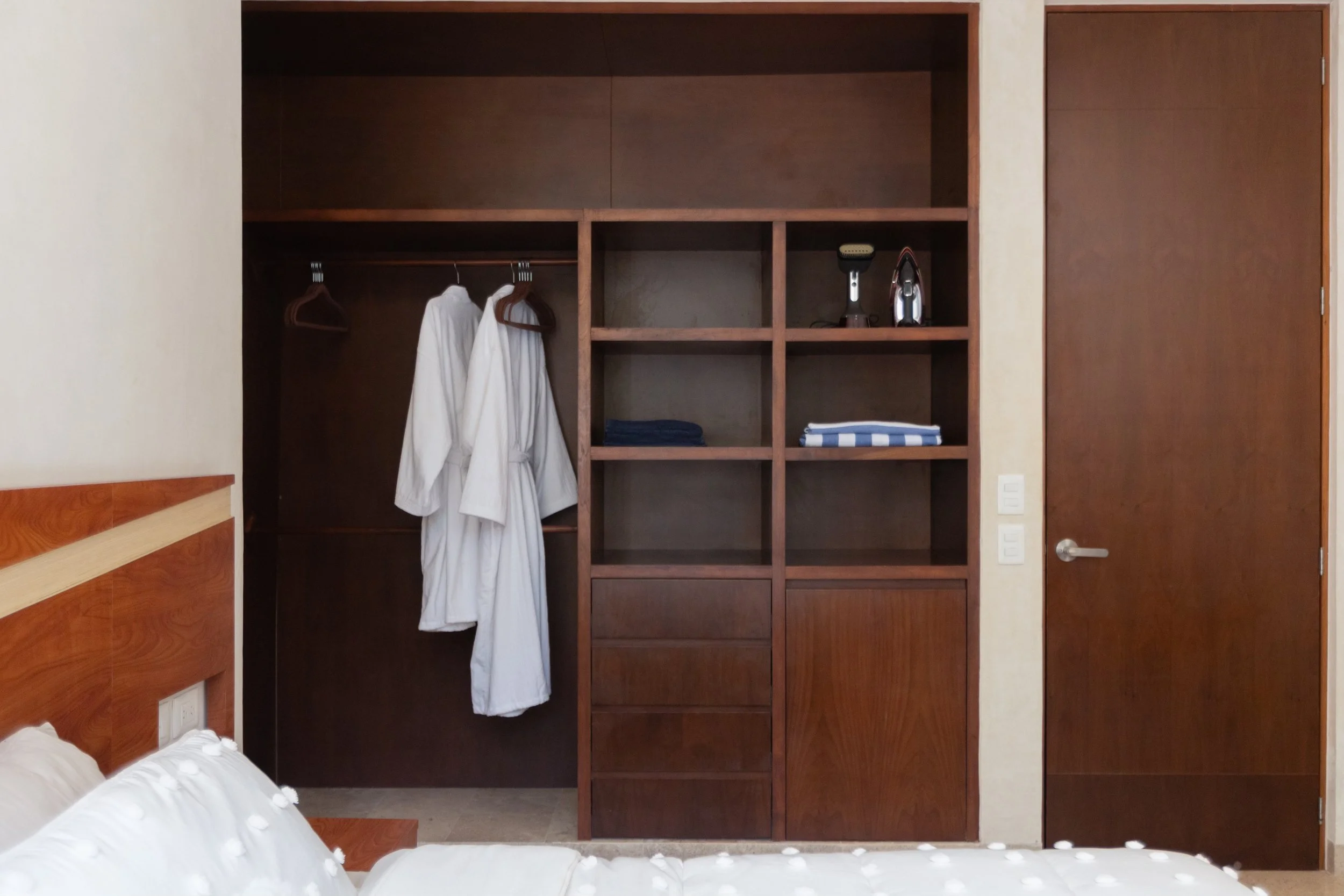 Wooden open closet with hanging white bathrobes, shelves with folded towels, a small iron, and an electric shaver in a bedroom.