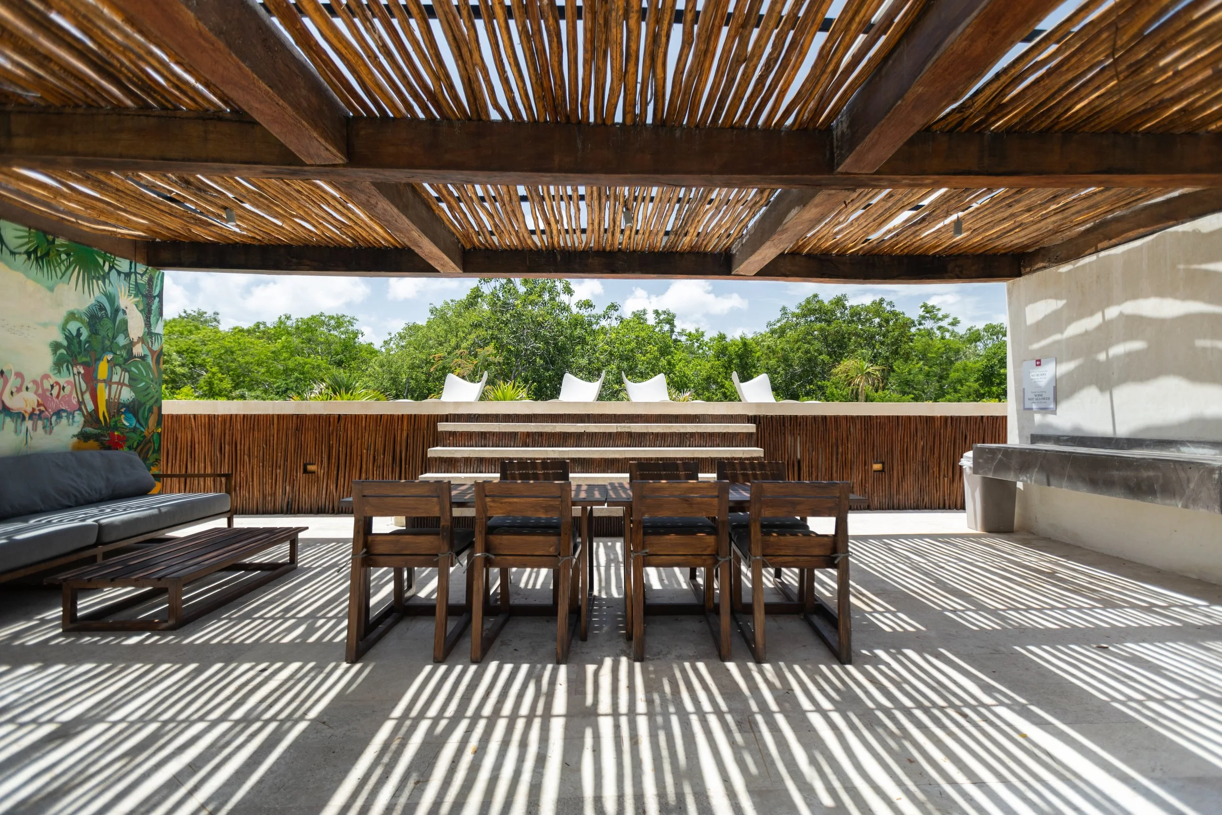 Outdoor patio with wooden furniture, including a table with eight chairs, a black couch, and a long counter on the right. The ceiling has a wooden slat design, casting striped shadows on the concrete floor. In the background, lush green trees and a partly cloudy sky are visible.