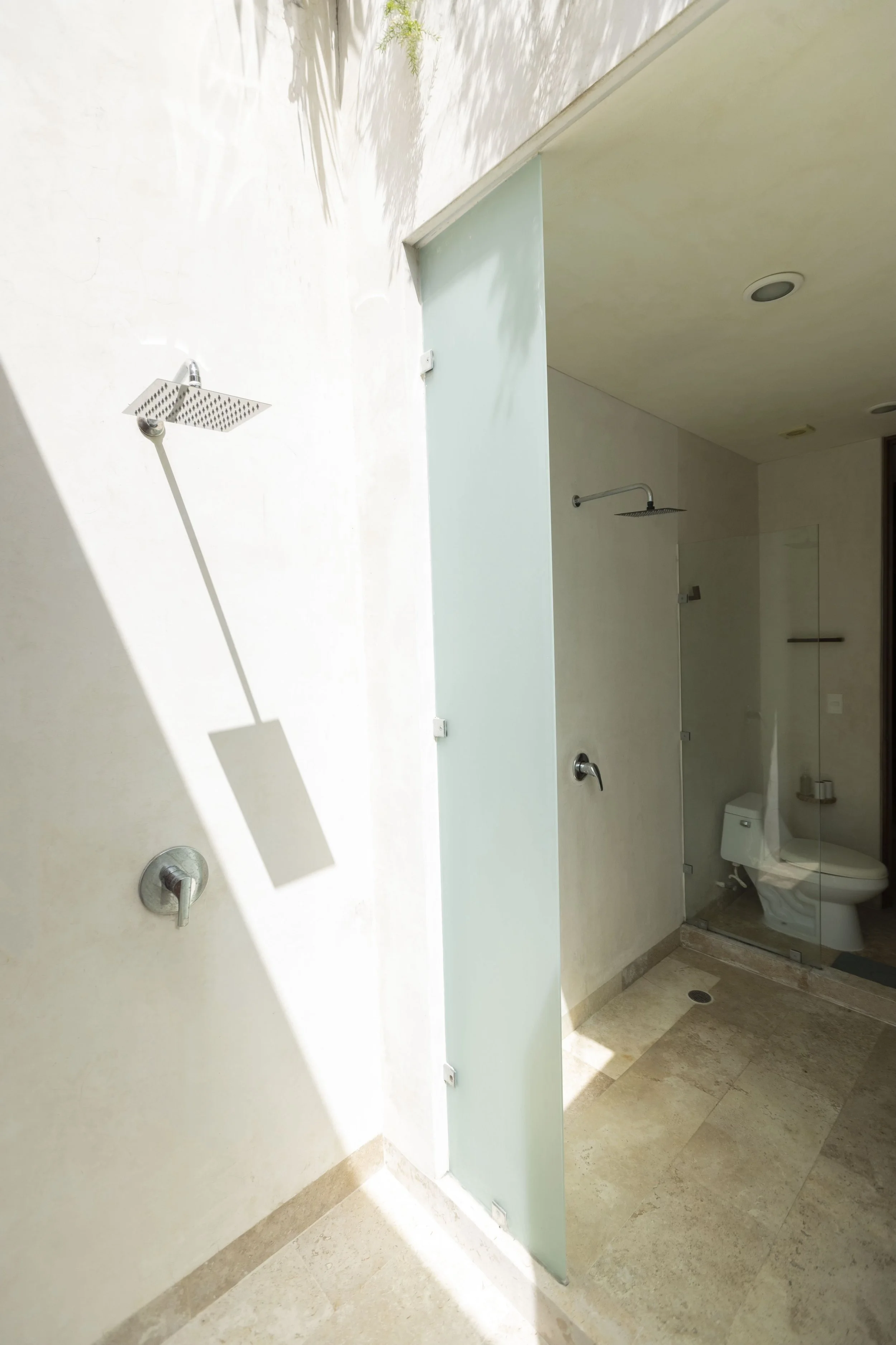 A modern outdoor shower area with a rainfall showerhead on a wall, a glass panel for privacy, and a view of the bathroom with a toilet inside.