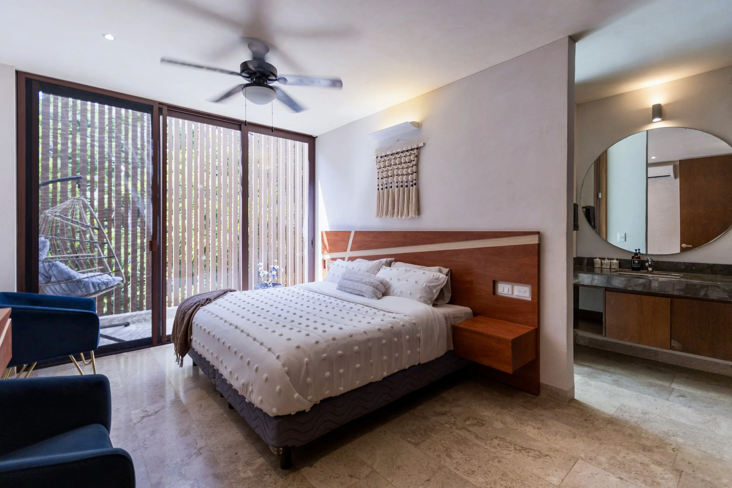 Modern hotel room with a bed, sliding glass door with vertical blinds, balcony with hanging chair, ceiling fan, and bathroom with round mirror and sink.