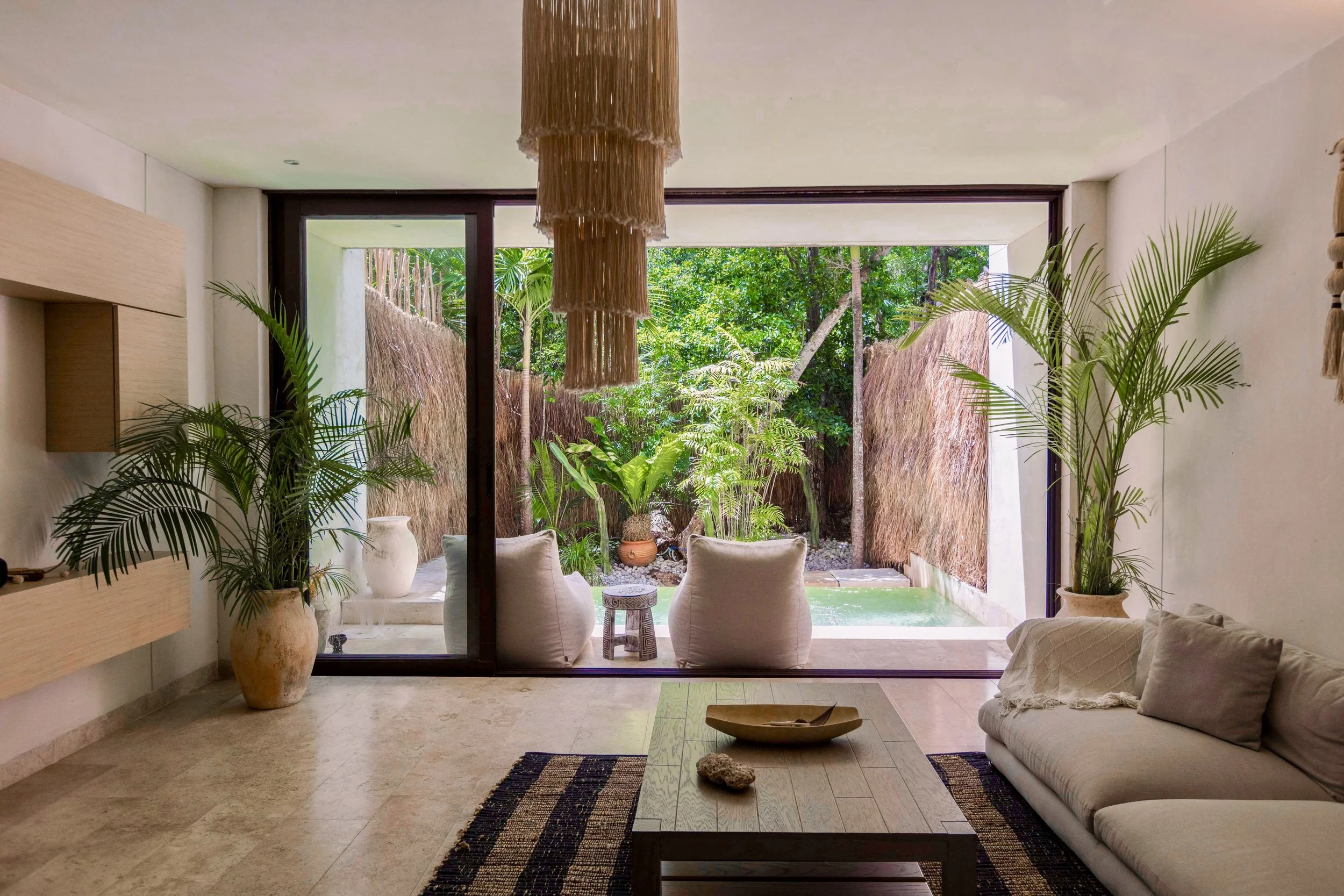 Living room with sliding glass door opening to a lush garden with tropical plants and a pool, decorated with potted plants, a beige sofa and a wooden coffee table.