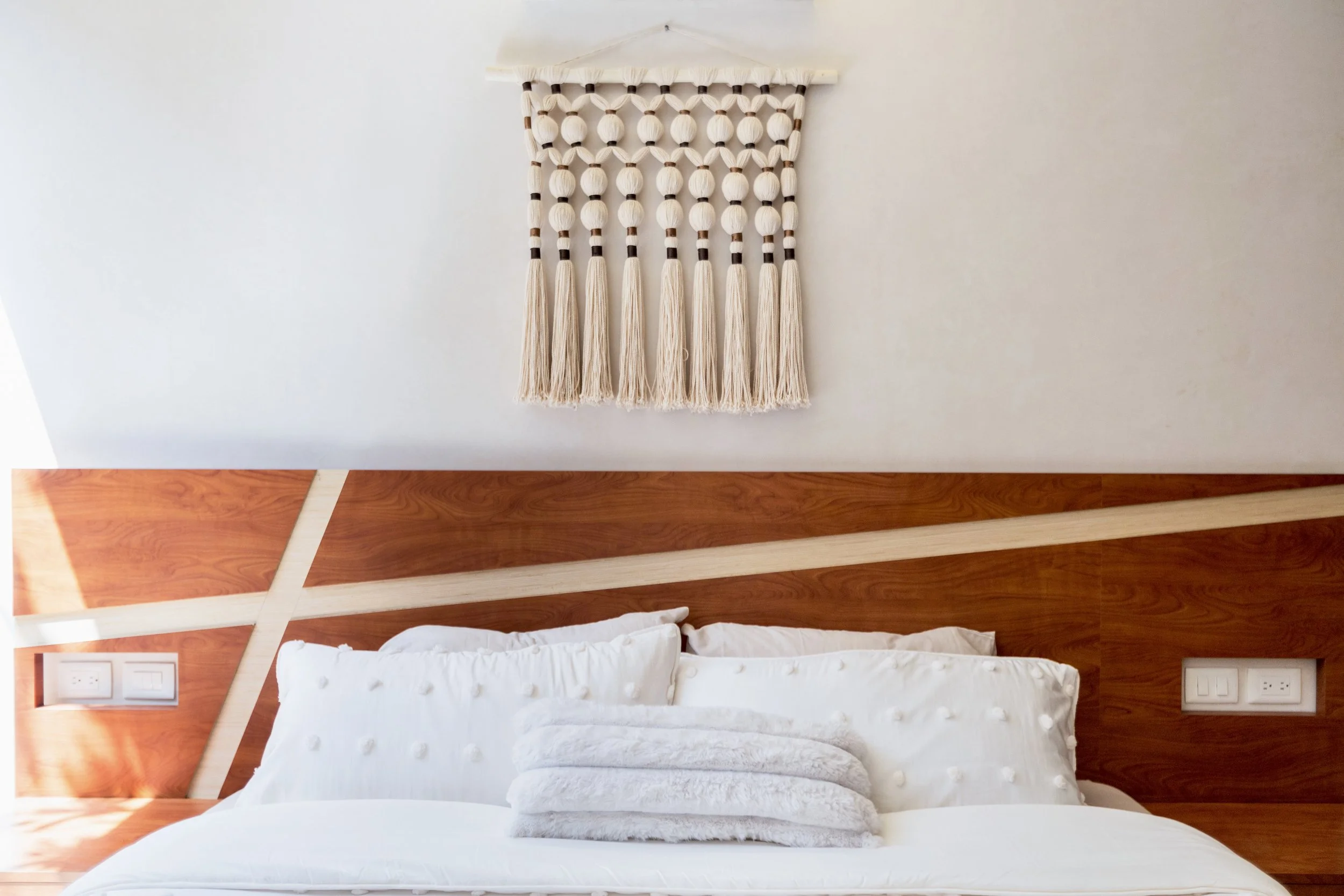 A bedroom with a white bed, decorated with white pillows, and a wooden headboard with a diagonal design; a bohemian-style wall hanging with tassels and beads above the bed; electric outlets and switches on the walls.