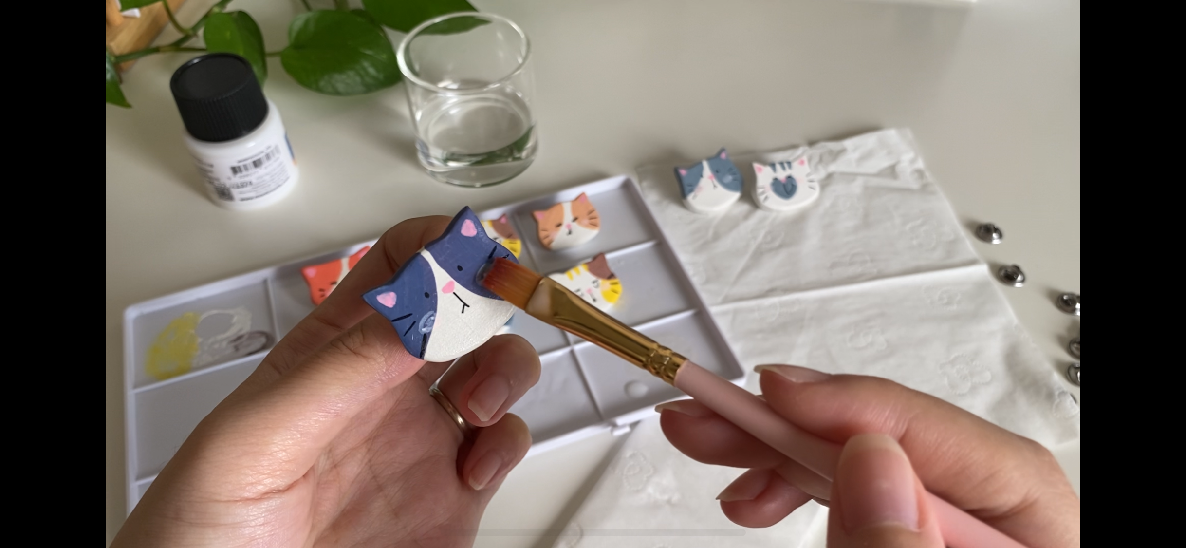 Person painting a decorative animal-shaped object with a small paintbrush, surrounded by similar painted objects on a table, with art supplies and a glass of water in the background.