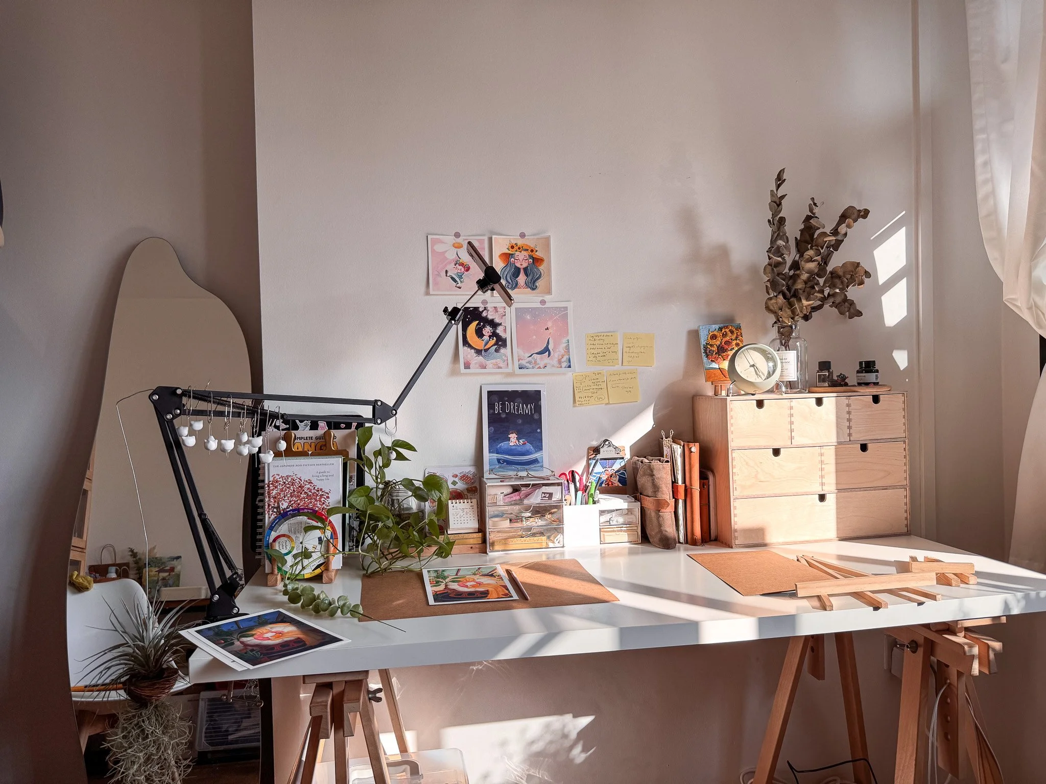 Sunlit workspace with art supplies, plants, artwork, and a wooden drawer on a white desk.