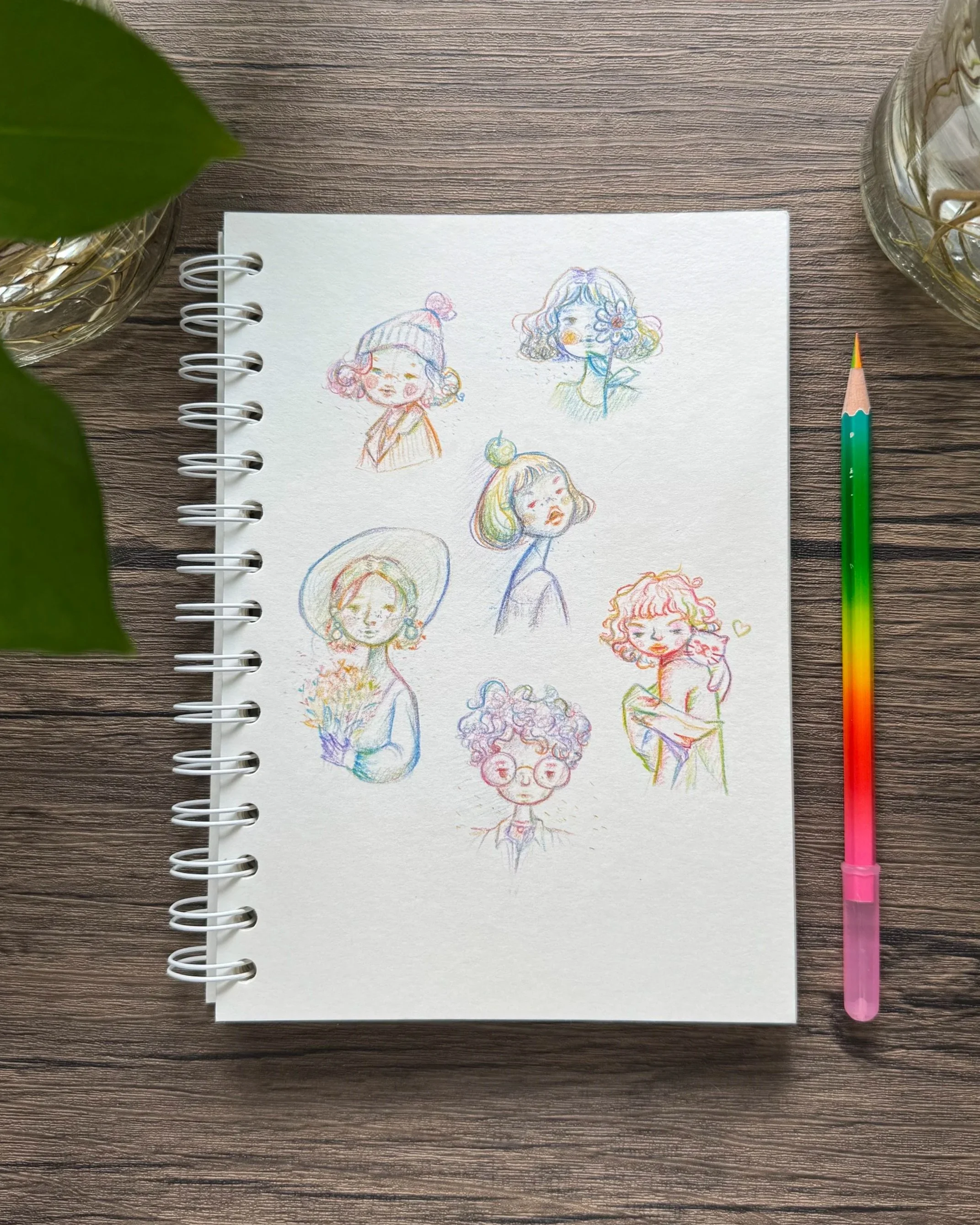Colorful sketches of various girls with different hairstyles and accessories, drawn on a white spiral notebook on a wooden table, with a multi-colored pen placed beside it.
