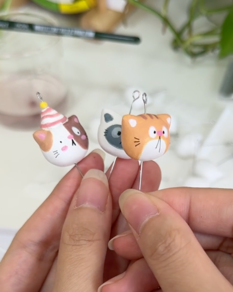 Three handmade cat figurines, each with a wire loop on top, held between fingers. One has a party hat, one is a gray and white, and one is orange with stripes.
