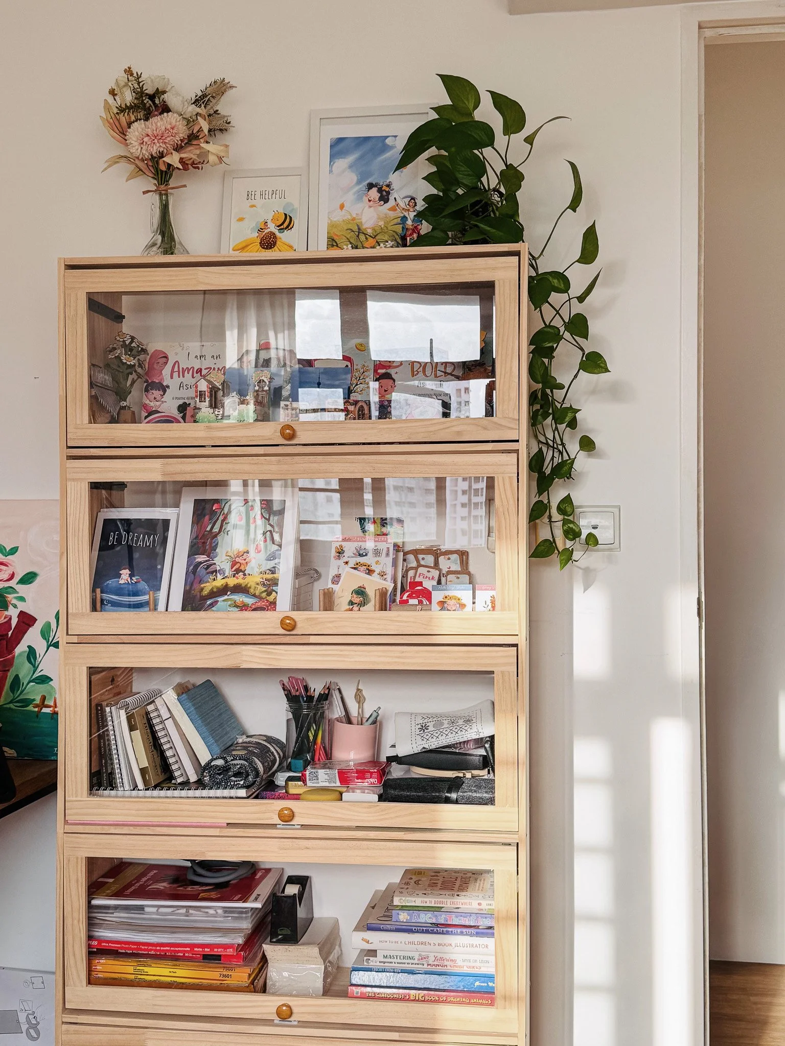 A wooden shelf with four sections containing books, stationery, and decorative items, along with framed artwork and a plant on top.