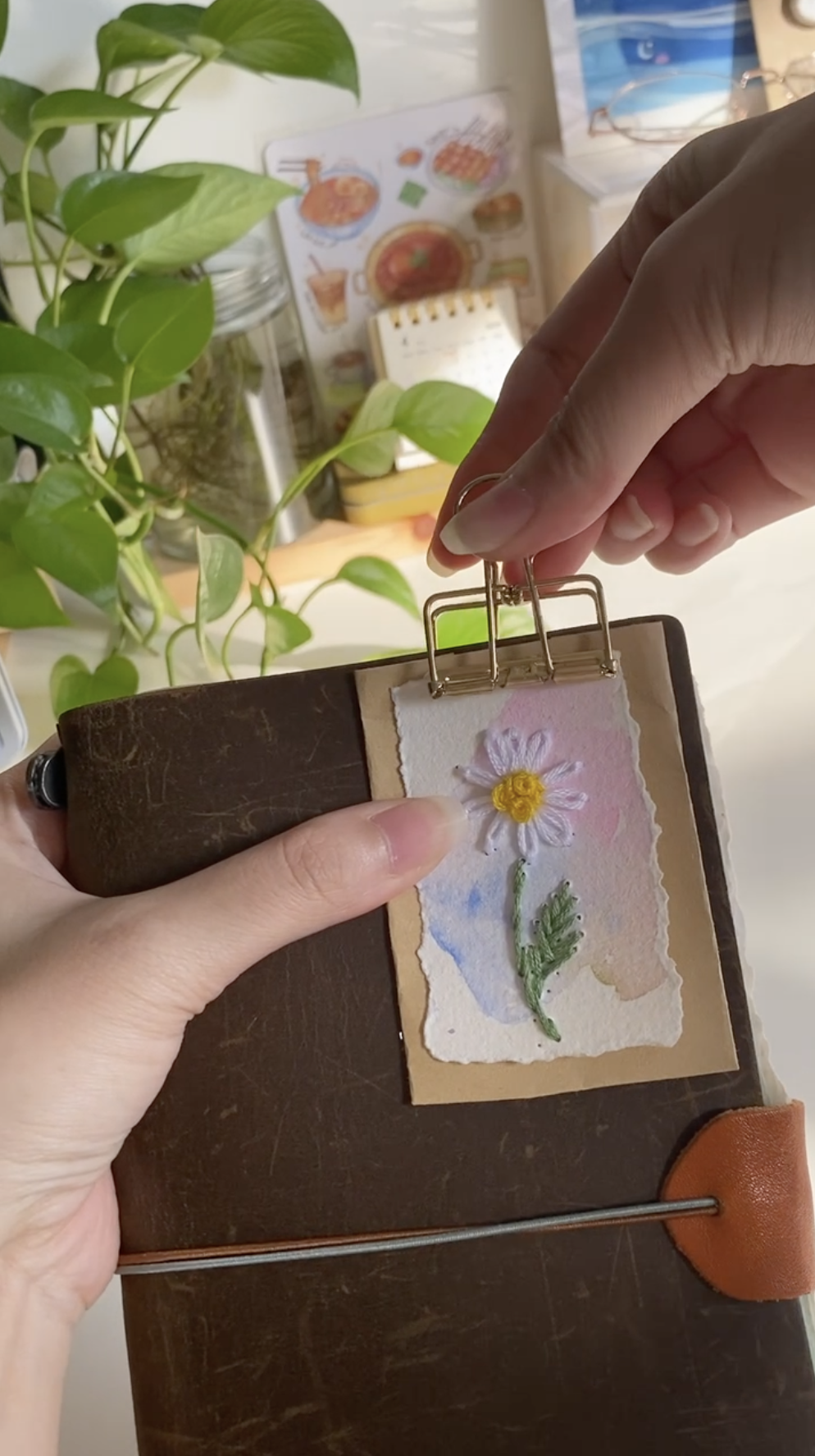 Person holding a small leather-bound journal with a decorative paper and embroidered flower ornament attached on the front cover.