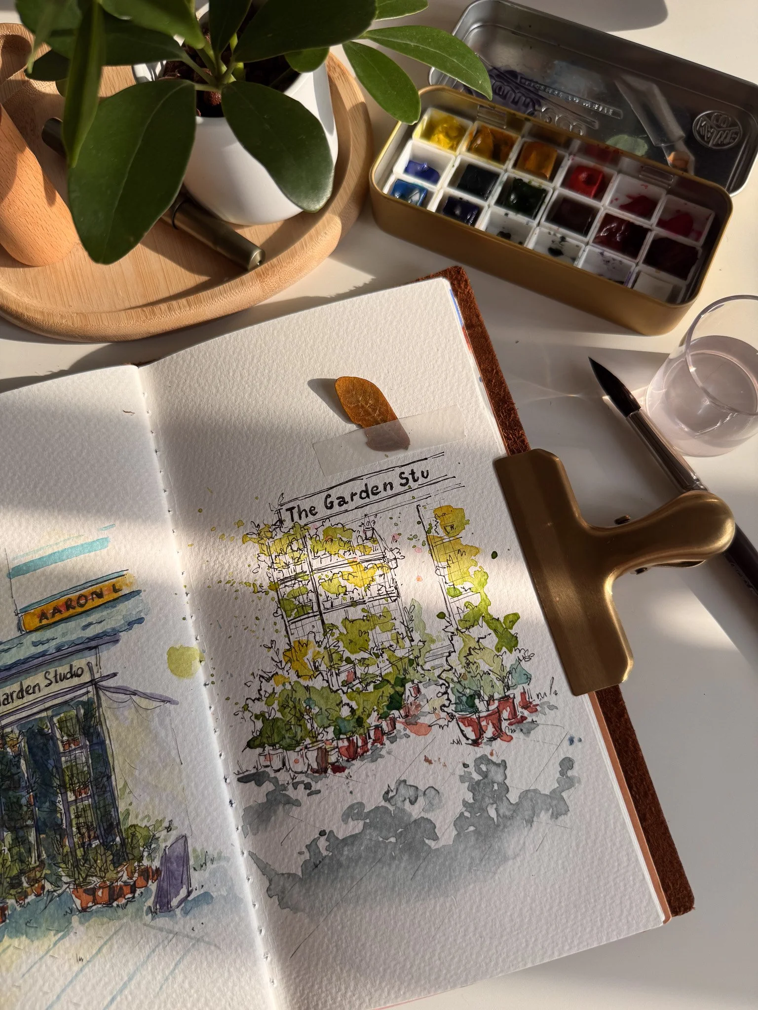 A sketchbook displaying a watercolor painting of a plant-covered structure labeled 'The Garden Stu', with a potted plant, watercolor palette, pen, and glass of water on a white table.