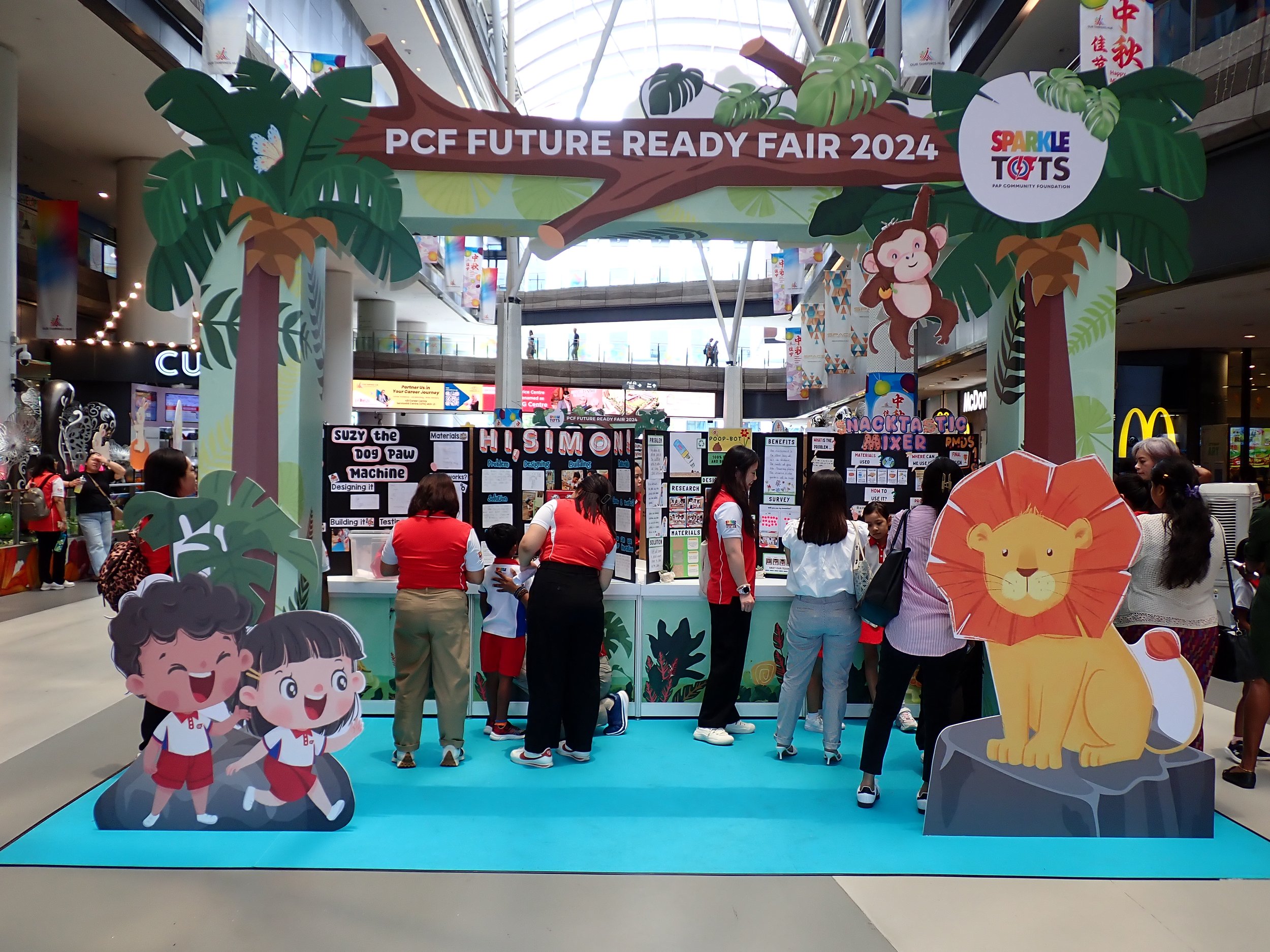 Children and adults gather at a display booth for the PCF Future Ready Fair 2024 in a shopping mall. The booth features cartoon jungle animals, including a lion and a monkey, with decorative trees and a large sign overhead. Various informational posters and interactive activities are visible.