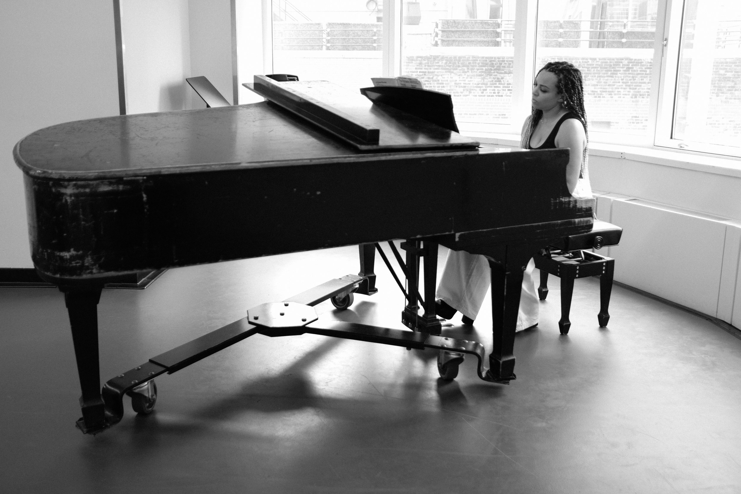 A woman playing a grand piano in a room with large windows, black and white photo.