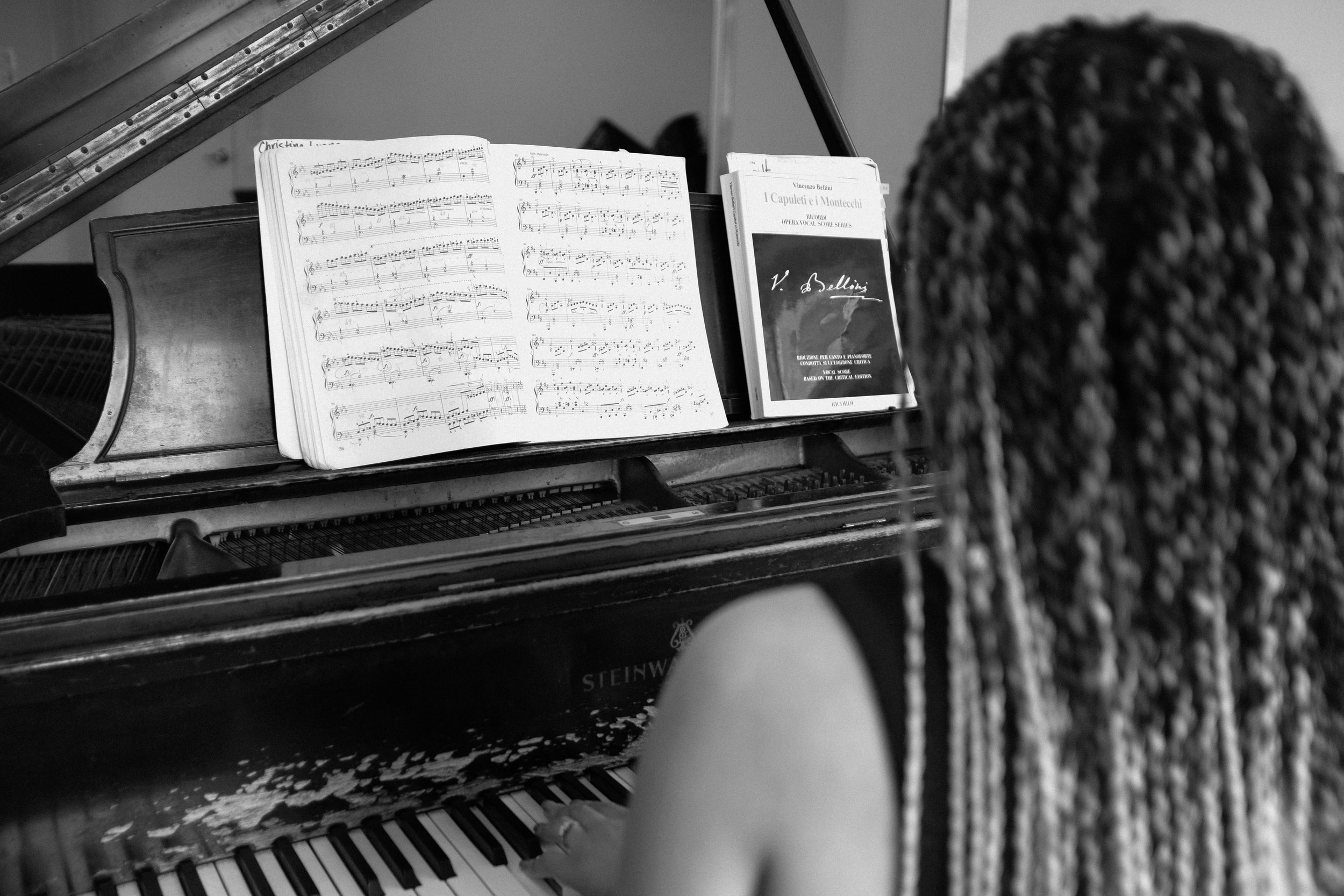 A person with long braided hair playing a Steinway grand piano with sheet music and a music book on the music stand.