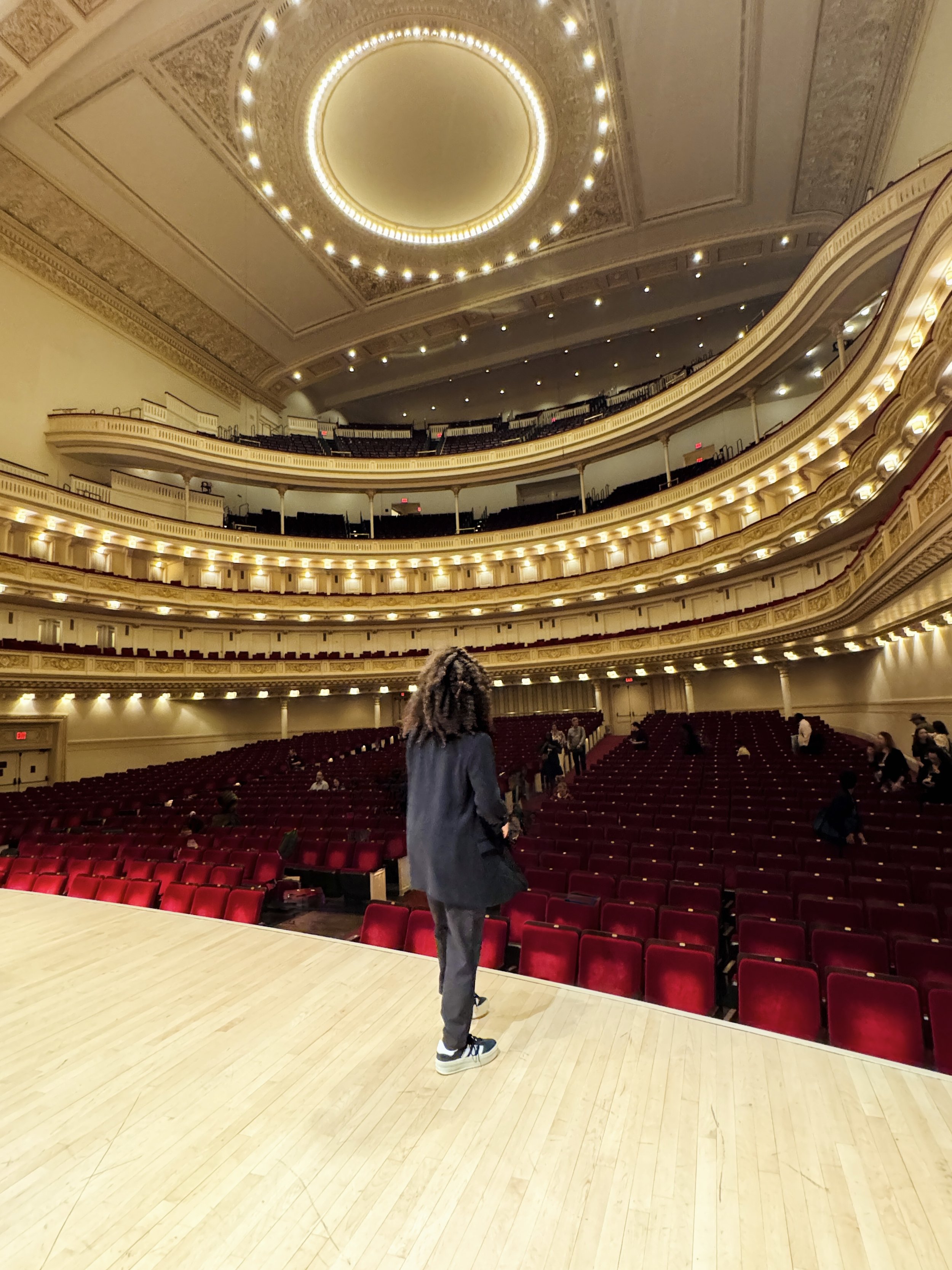 Person standing on the stage at Carnegie Hall with ornate balconies and red seats.