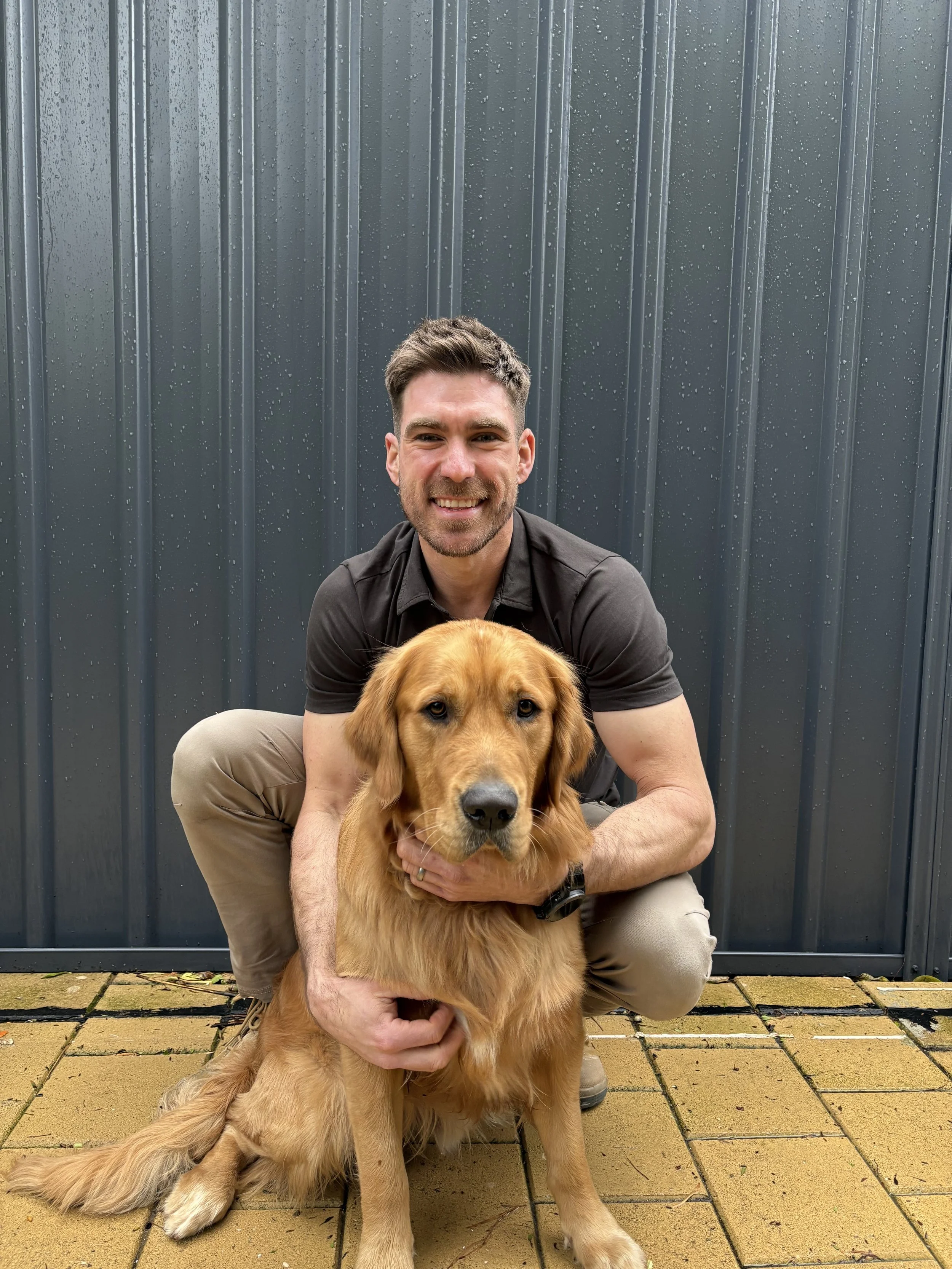 A man with short dark hair squatting on a yellow brick patio, holding a golden retriever, with a dark gray metal fence covered in raindrops in the background.