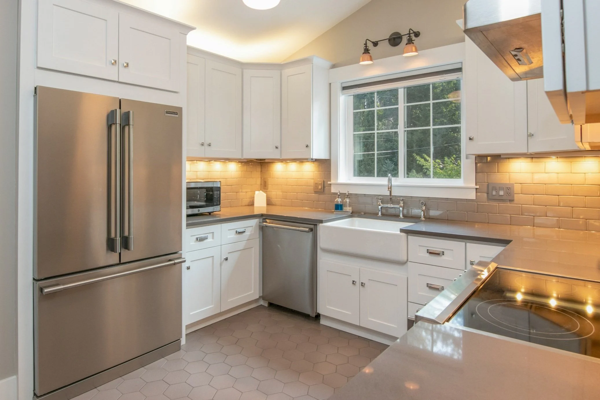Modern kitchen with white cabinets, stainless steel refrigerator, microwave, countertop, window, and under-cabinet lighting.