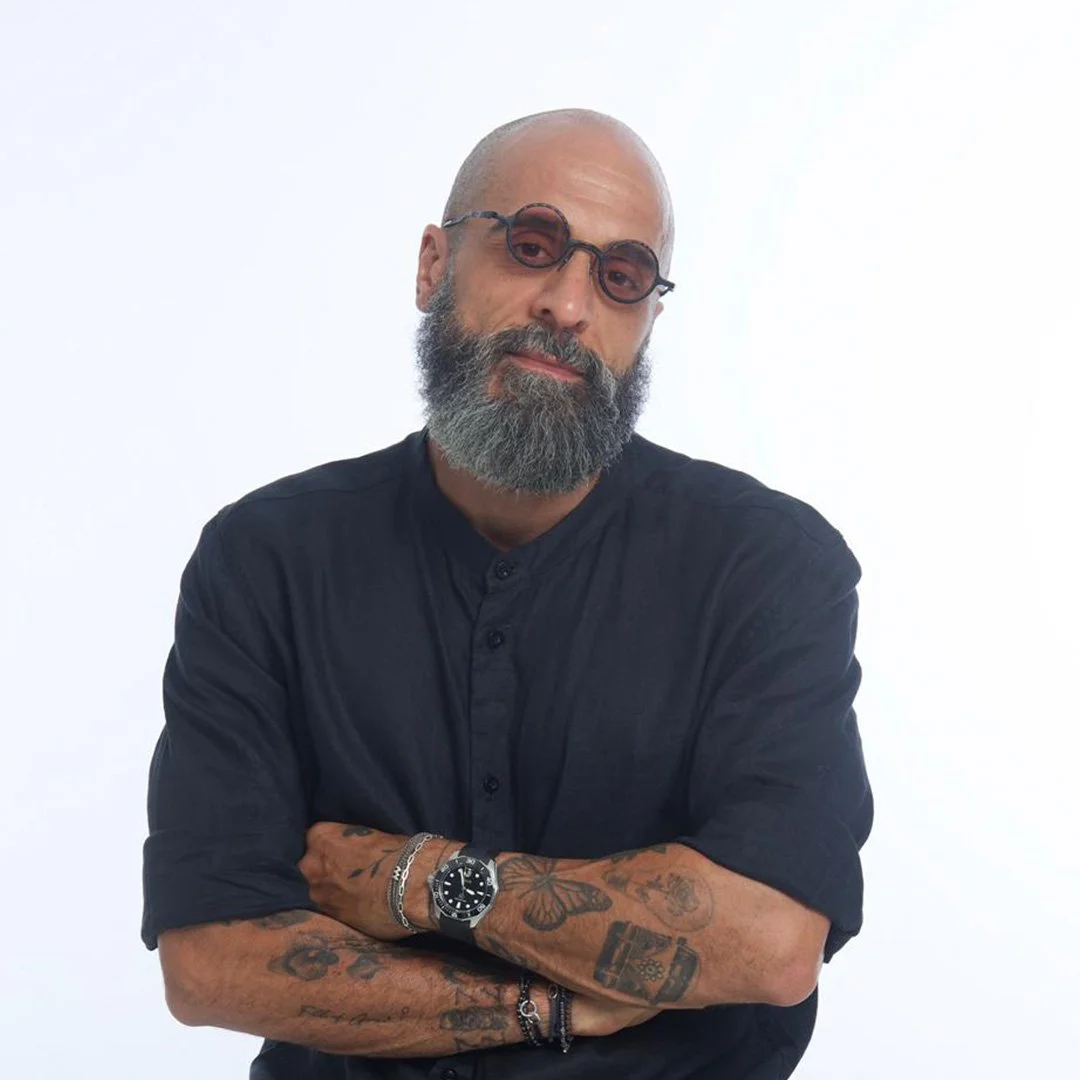 Man with a beard, tattooed arms, wearing black shirt, watch, and glasses, crossing arms, standing against a white background.