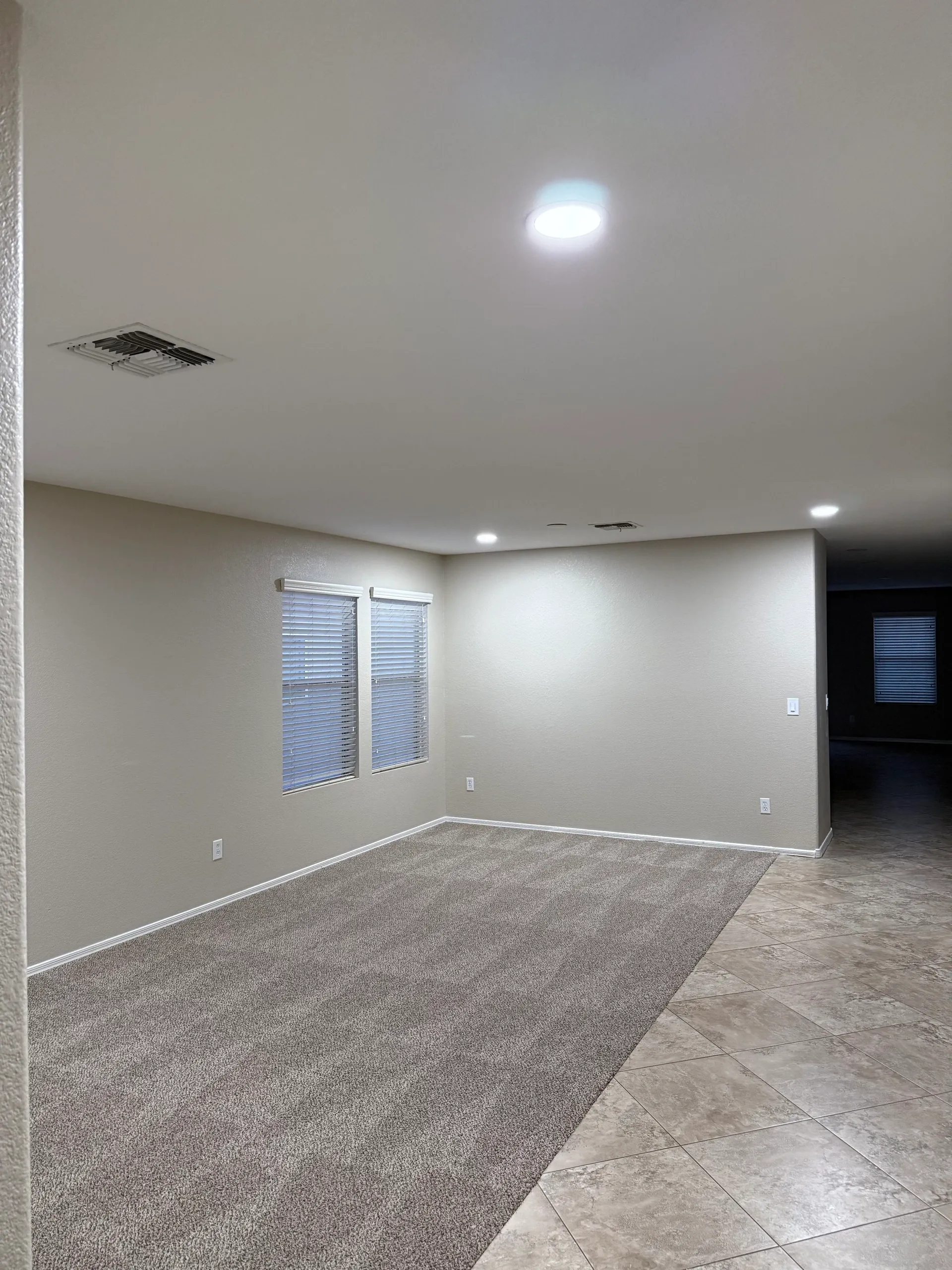 Empty living room with beige walls, two windows with blinds, carpeted floor, and tile flooring near the entrance, illuminated by ceiling lights.