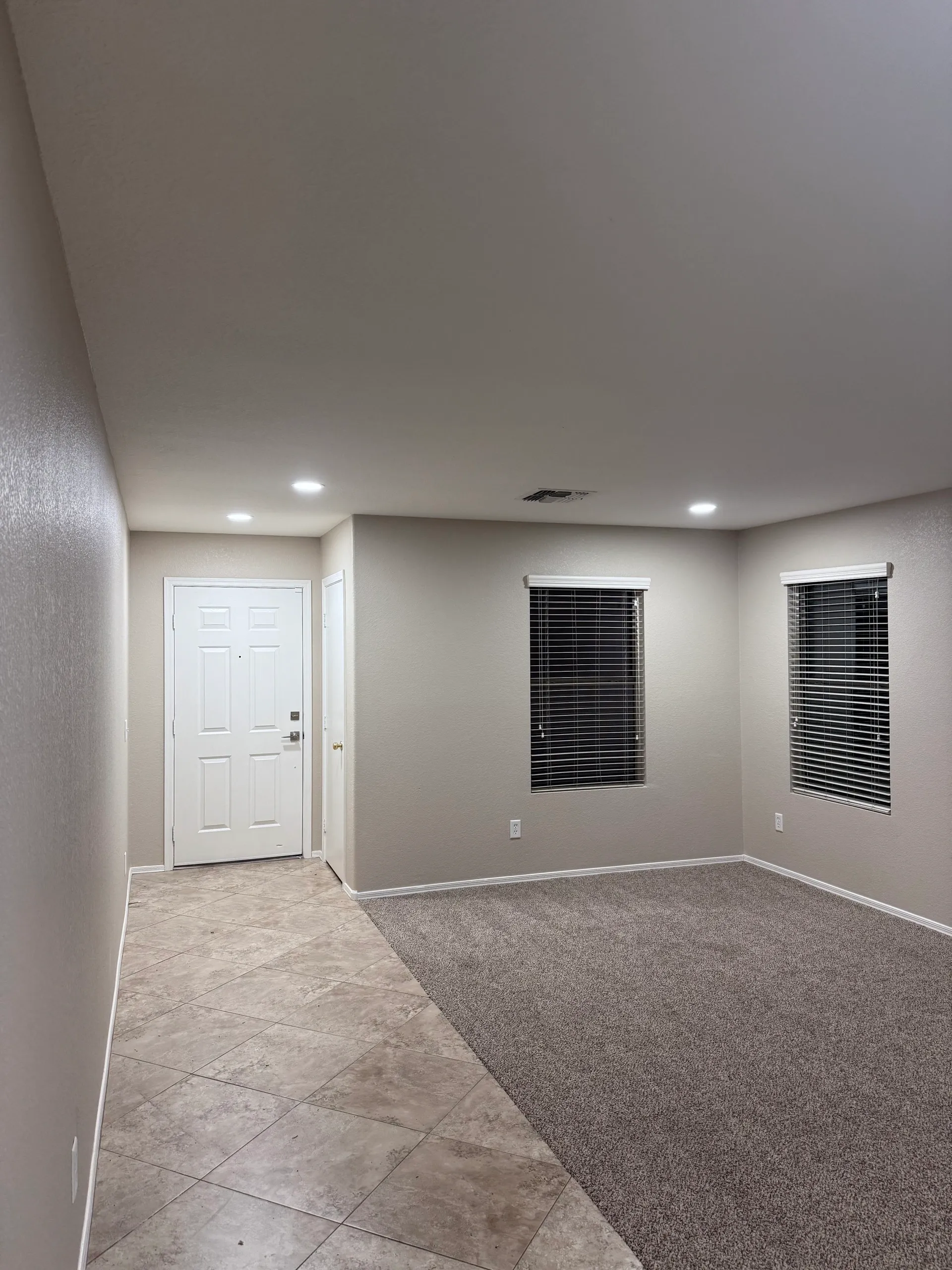 Empty living room with tiled entrance area, beige carpeted floor, beige walls, and two windows with closed horizontal blinds.