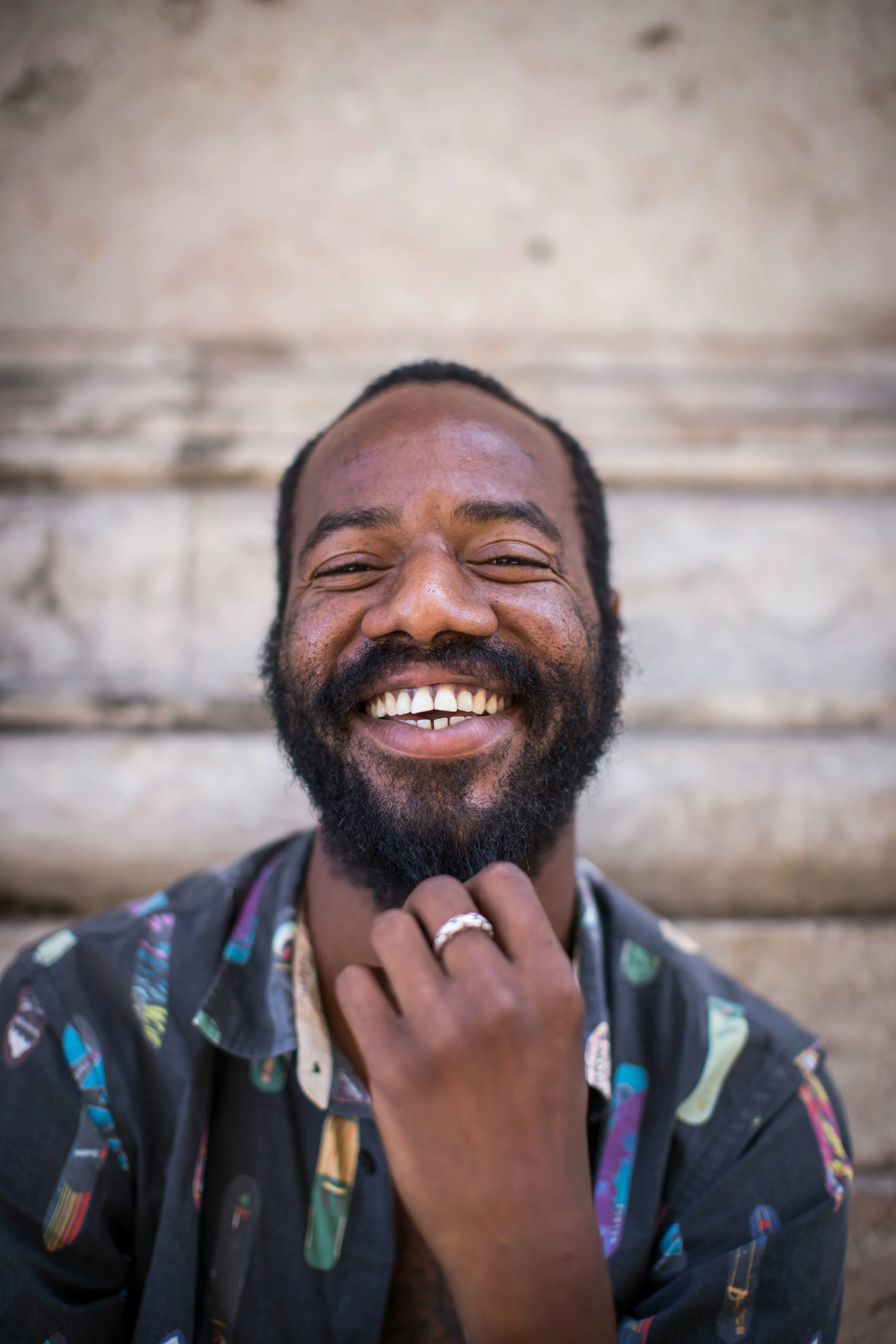 A man with a beard and mustache smiling, touching his chin with his hand, wearing a colorful shirt, standing in front of a wooden background.