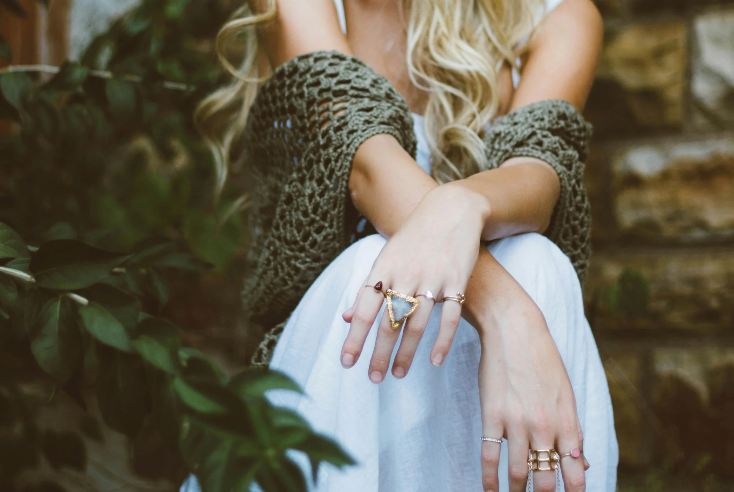 A person resting their arms on their knees, wearing multiple rings, with a large gemstone ring on their middle finger, in an outdoor setting with greenery and a wooden background.
