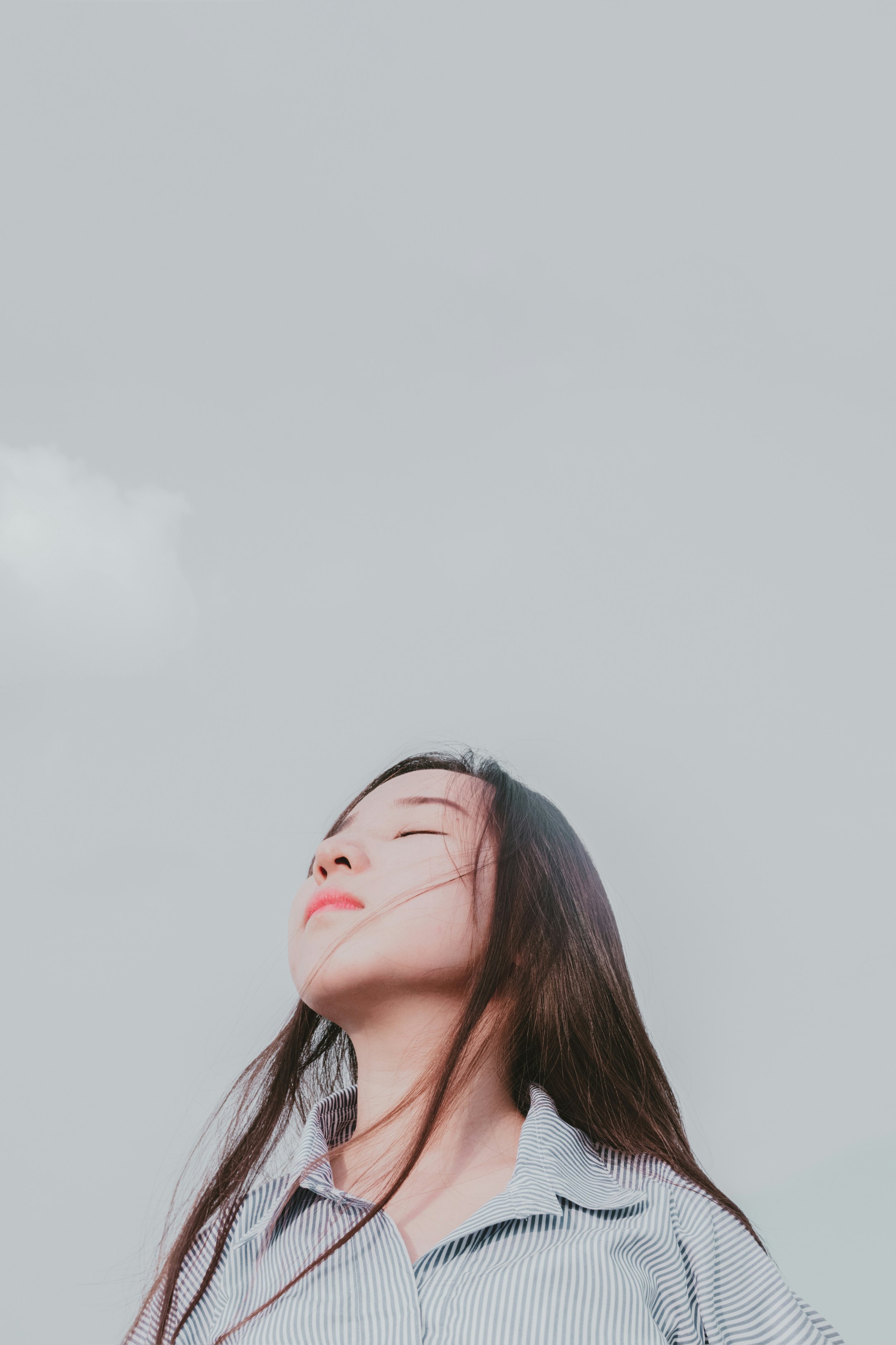 A woman with closed eyes and wind in her hair stands outdoors against a cloudy sky.