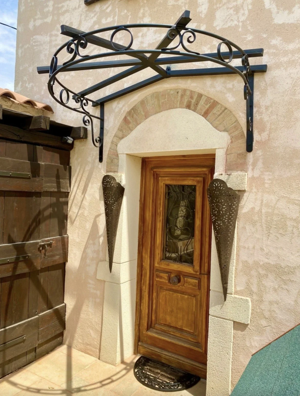 Porte en bois avec une arche en briques au-dessus, décorée avec des lampes métalliques coniques et un support en métal ornementé au-dessus, dans une façade de maison en pierre beige.