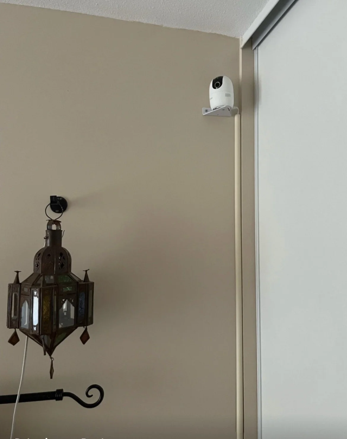 Caméra de sécurité blanche montée au coin du mur beige avec un support métallique, et un lustre marocain en métal et verre suspendu, avec une barre en fer noir.