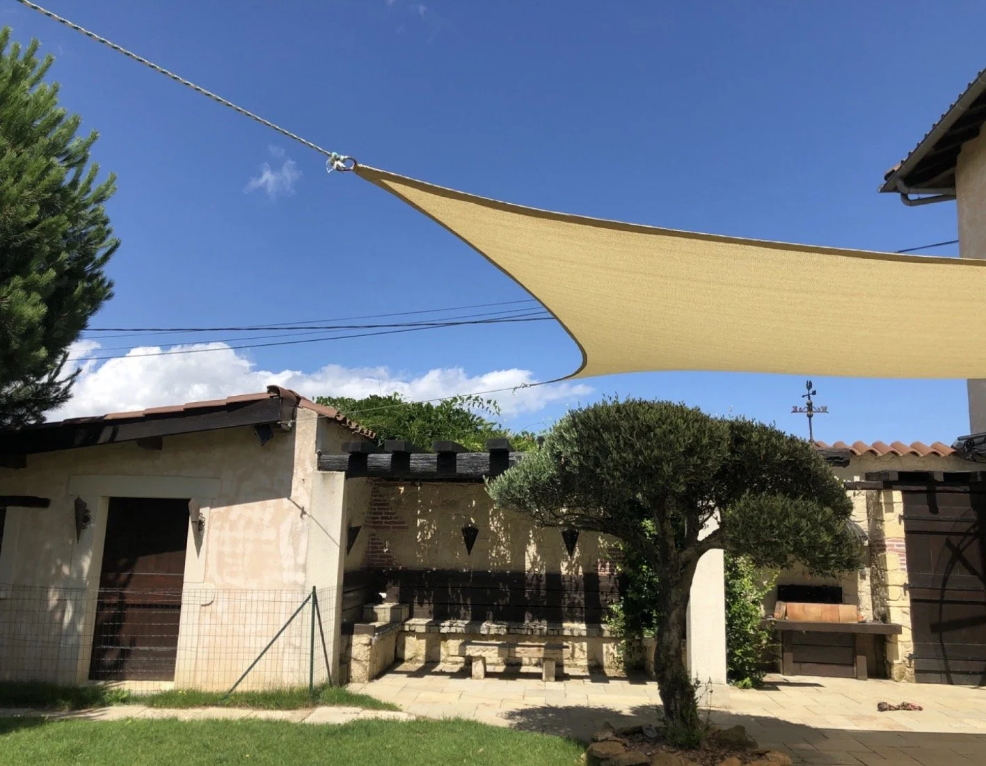 Jardin avec arbre, petite construction, voiles d'ombrage beige en haut, ciel bleu et nuages.