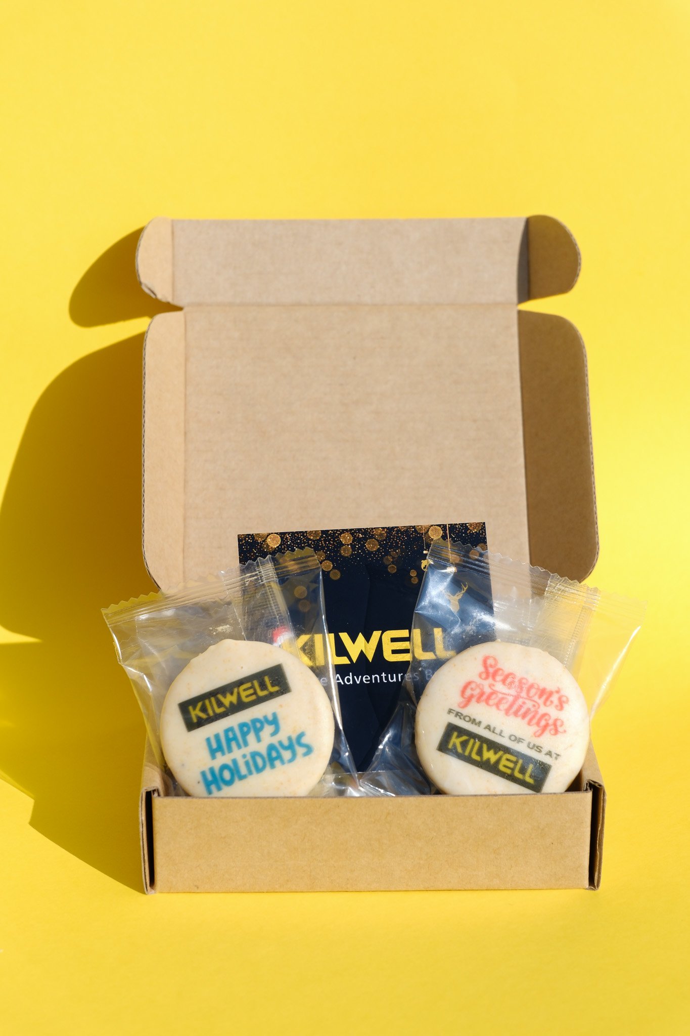 Open cardboard box with two decorated cookies inside, one with 'HAPPY HOLIDAYS' and the other with 'Season's Greetings', on a yellow background.