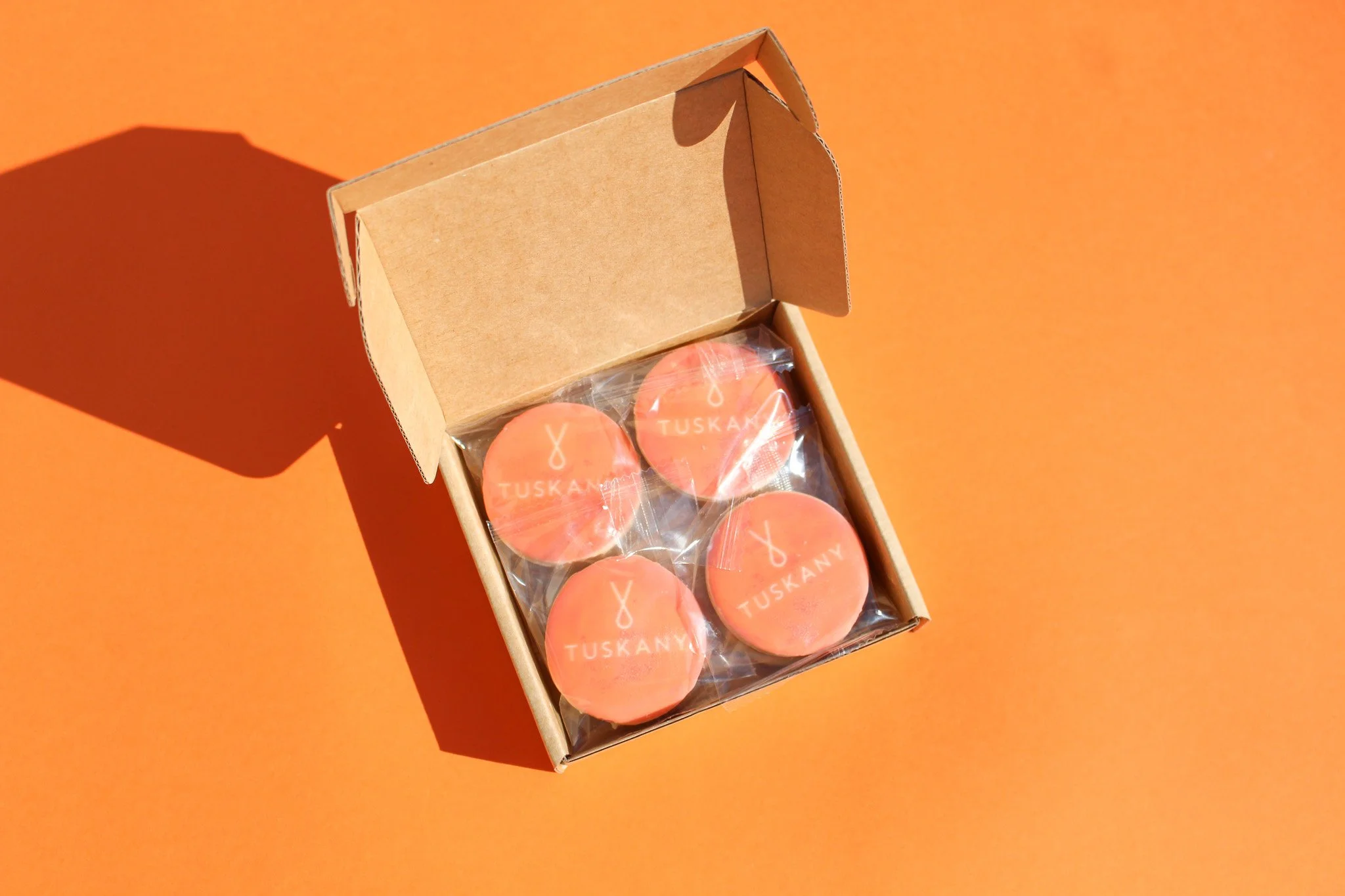 Open cardboard box containing four individually wrapped peach-colored candies with the word 'TUSKANY' on each.