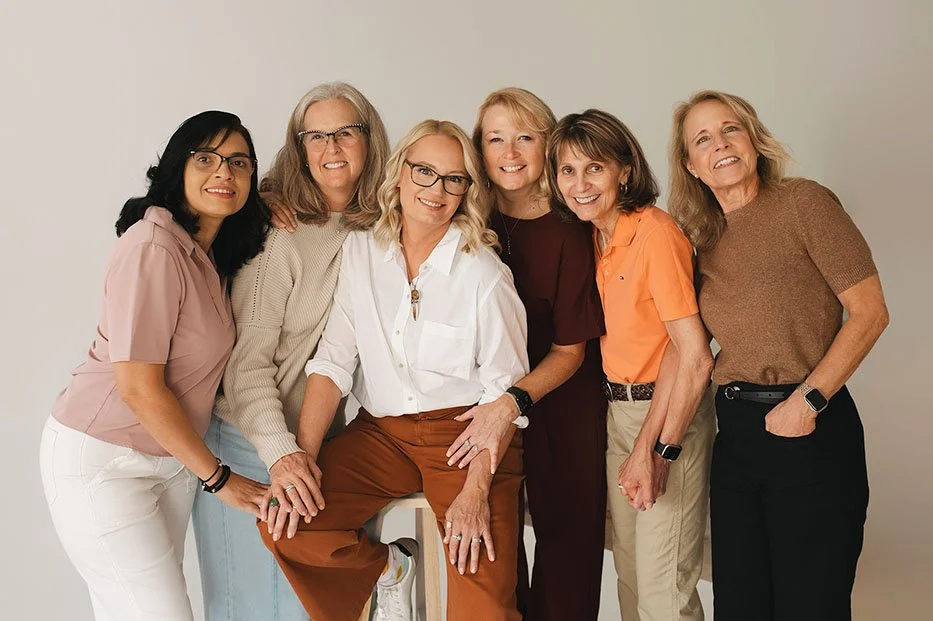Six women standing close together and smiling in front of a plain white background.