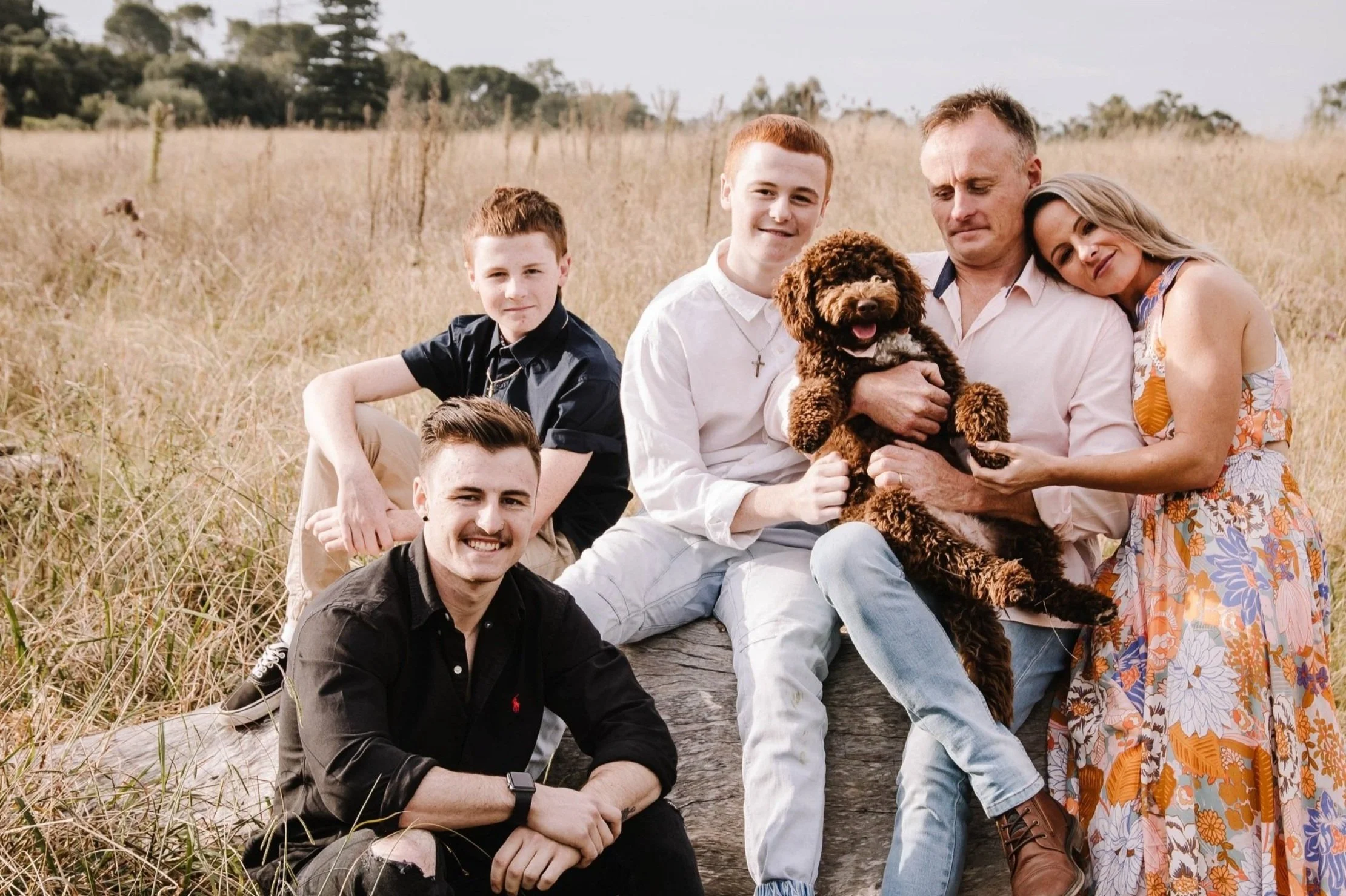 Family of six sitting outdoors in a field with dry grass, including a dog, smiling at the camera.