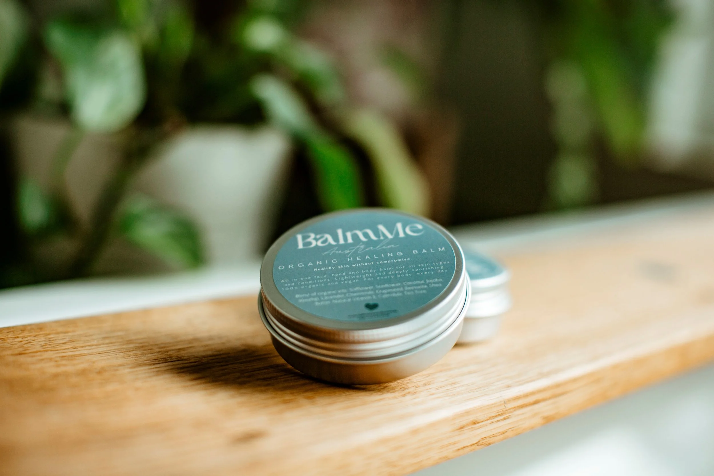 A tin container of BalmMe Australia organic healing balm resting on a wooden surface, with a blurry green plant in the background.