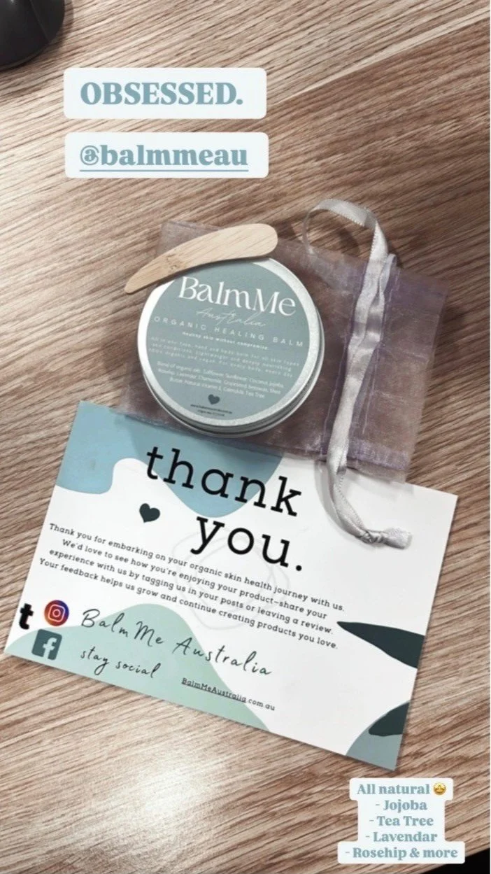 A promotional flyer and a small container of organic healing balm from Balm Me Australia on a wooden surface. The flyer thanks customers for their support and encourages social media engagement. The balm container has a wooden applicator on top and a clear pouch with a ribbon closure. The flyer mentions natural ingredients like jojoba, tea tree, lavender, rosehip, and others.