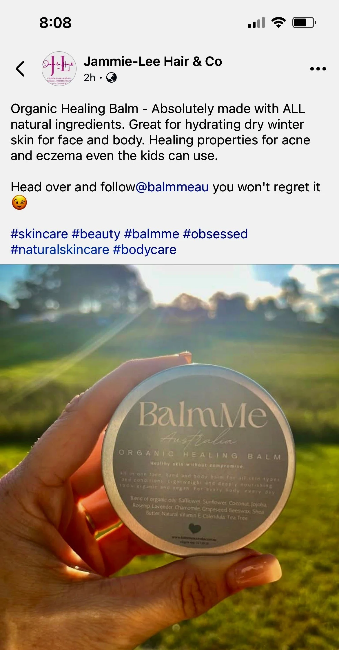 Hand holding a container of BalmMe Organic Healing Balm outdoors in a sunny field, with trees and a bright sky in the background.